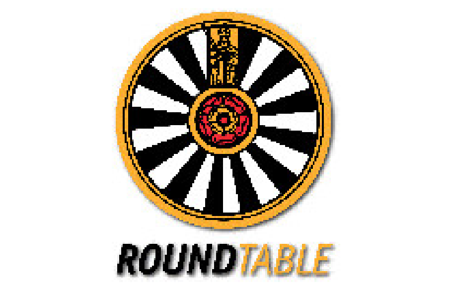 A circular sign with a black and white spiral pattern and a red rose in the center. The sign has a gold and black border. Below it, the text reads "ROUNDTABLE," with "ROUND" in black and "TABLE" in gold.