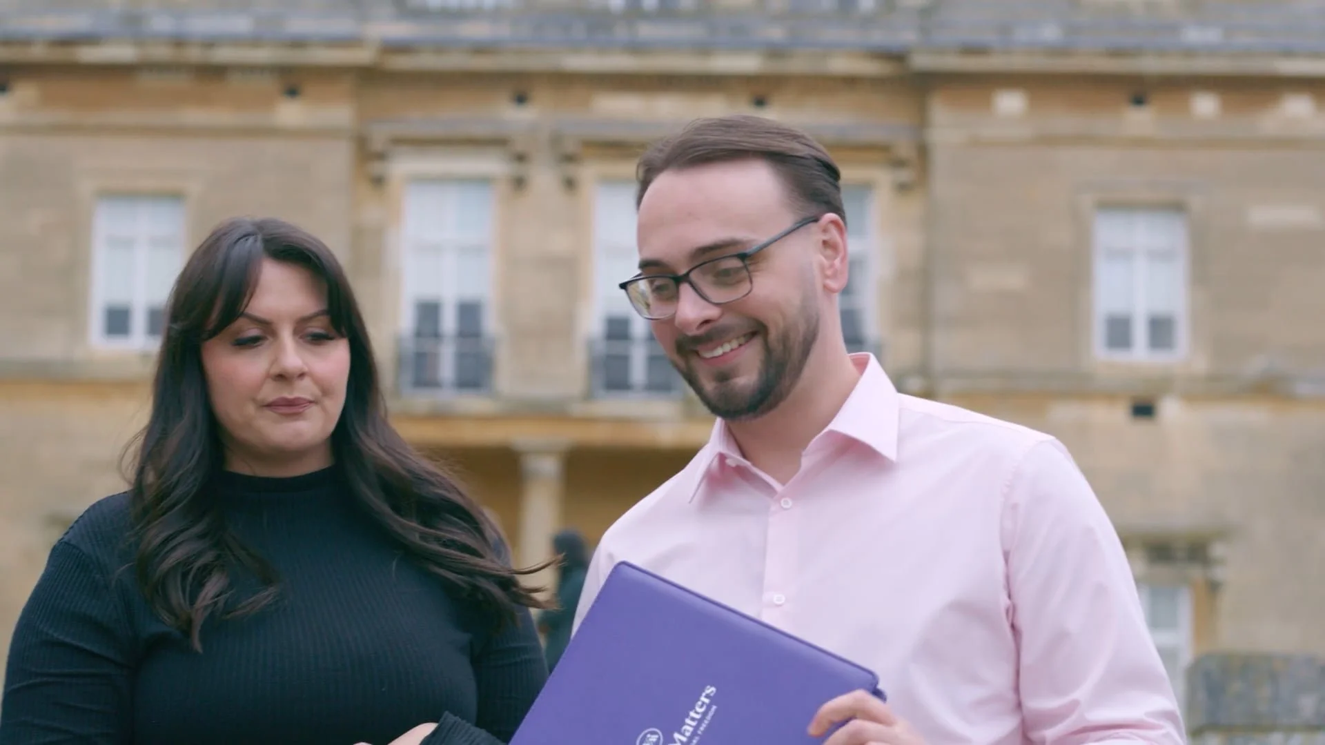 A man with glasses and a woman are outdoors looking at a purple folder with the word 'Matters' on it. They appear to be engaged in a conversation, with a historic stone building in the background.