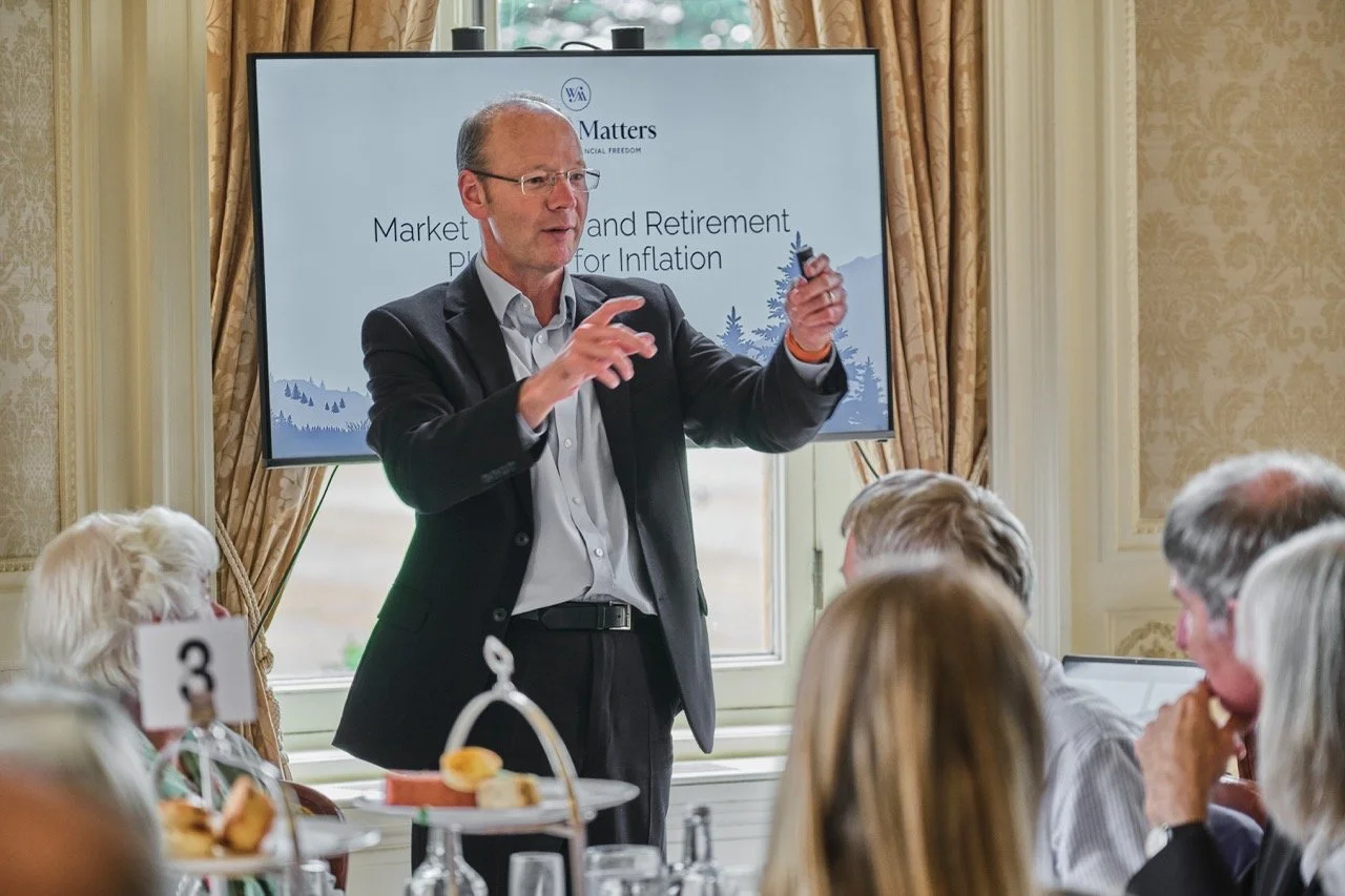 A man in a suit giving a presentation to an audience in a formal room with gold curtains and decorated walls. A large screen behind him displays the title 'Market and Retirement Planning for Inflation.' The audience is seated at tables with plates of food and numbered signs.