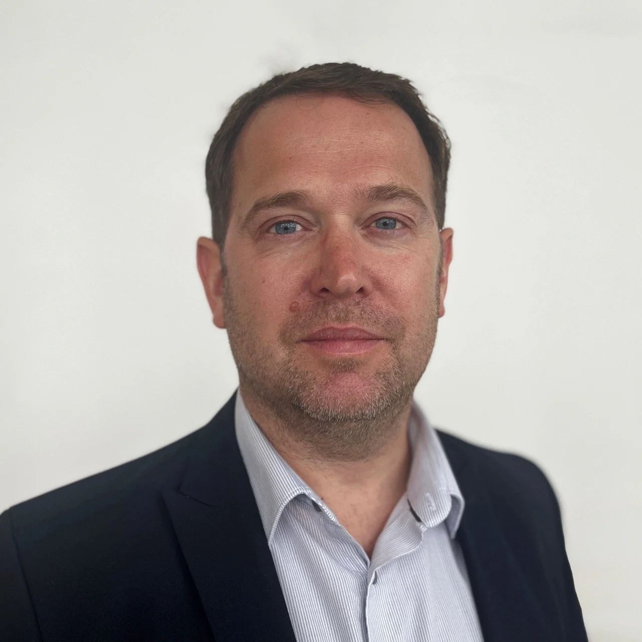 Professional headshot of a man with short brown hair, blue eyes, wearing a dark suit jacket and a collared shirt, standing against a plain white background.