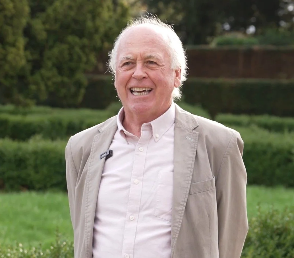 An elderly man with white hair smiling outdoors in a park with greenery in the background.