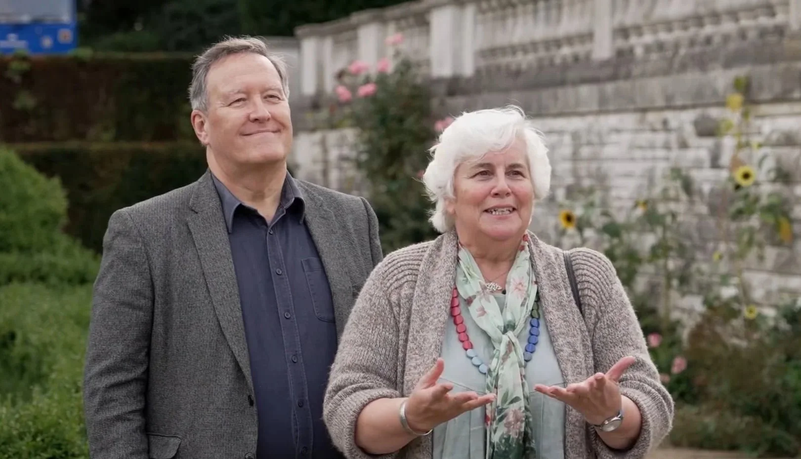 A man and an older woman stand outdoors with a garden and a brick wall in the background. The woman is speaking, gesturing with her hands, and the man is smiling.