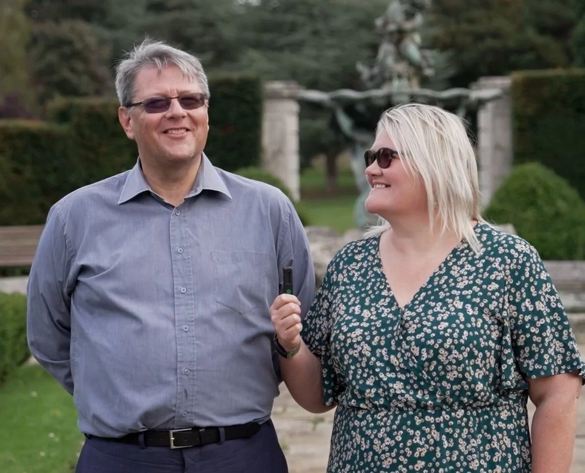 A man and woman smiling outdoors, with the woman holding a microphone towards the man. They are both wearing sunglasses and standing in front of a garden with statues and trees.