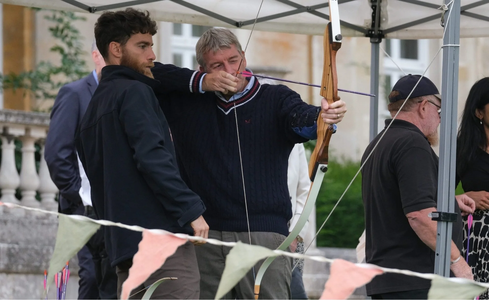 A man in a black sweater is aiming a bow and arrow, with another man standing next to him at an outdoor event.