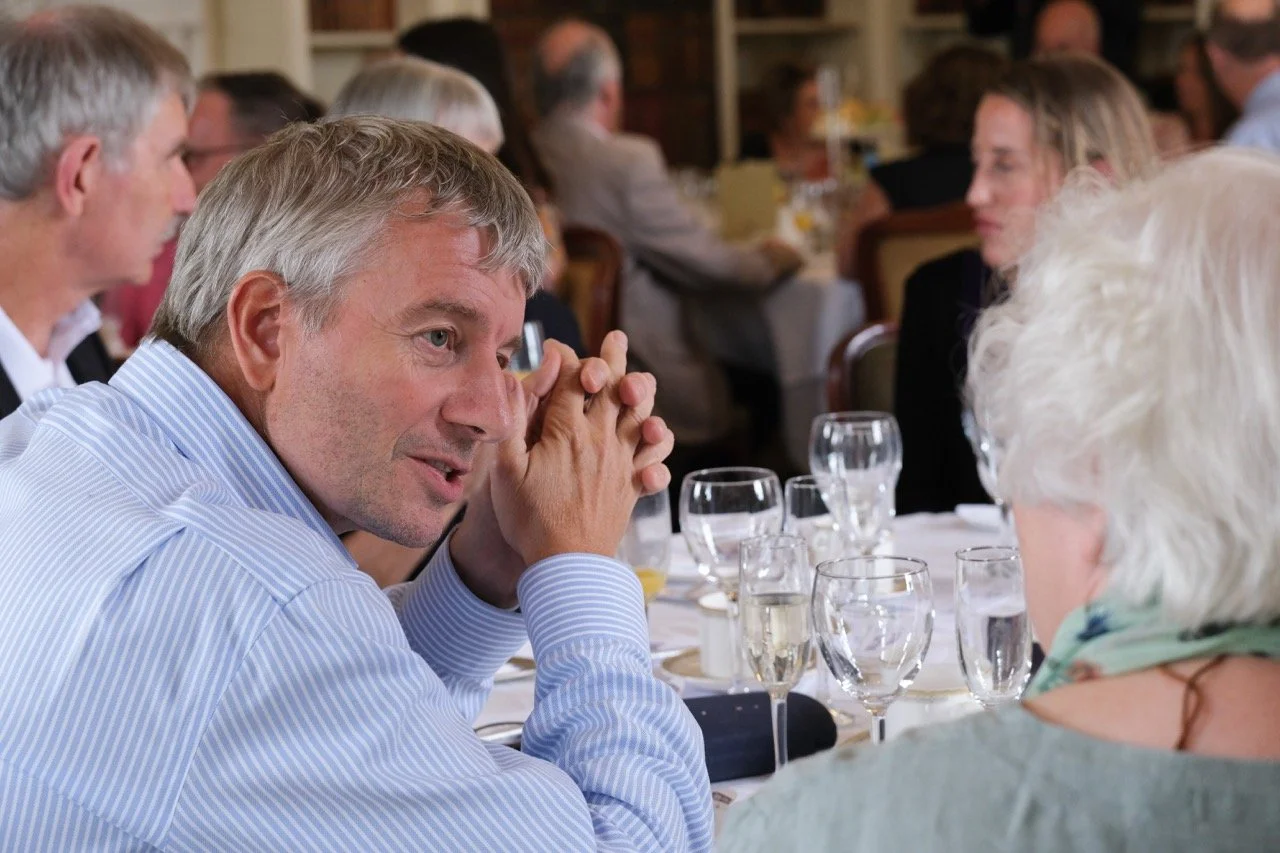 A man in a blue striped shirt is seated at a table with others, engaging in conversation in a formal dining setting with glasses and tableware.