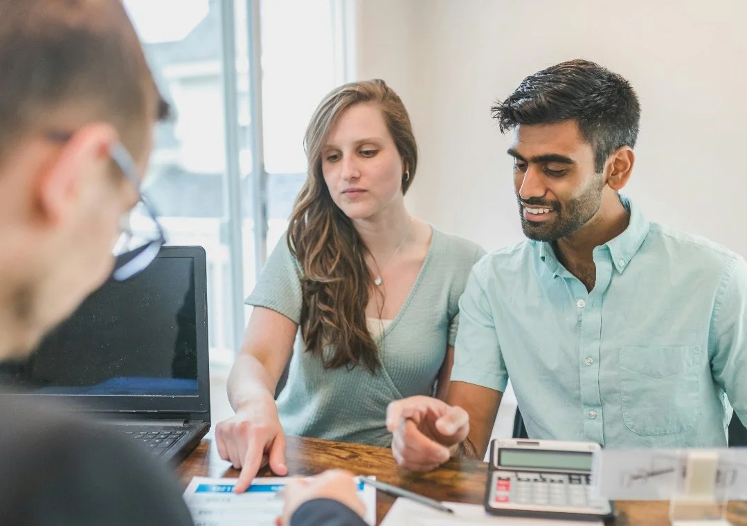 A woman and man sitting at a table with a third person whose face is blurred; the woman points to documents while the man smiles.