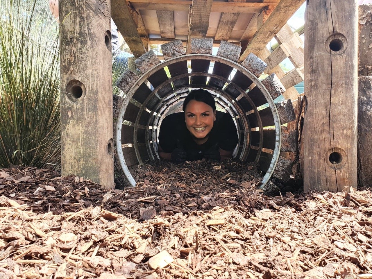 A woman smiling while crawling through a wooden and metal tunnel in an outdoor setting with wood chips on the ground.