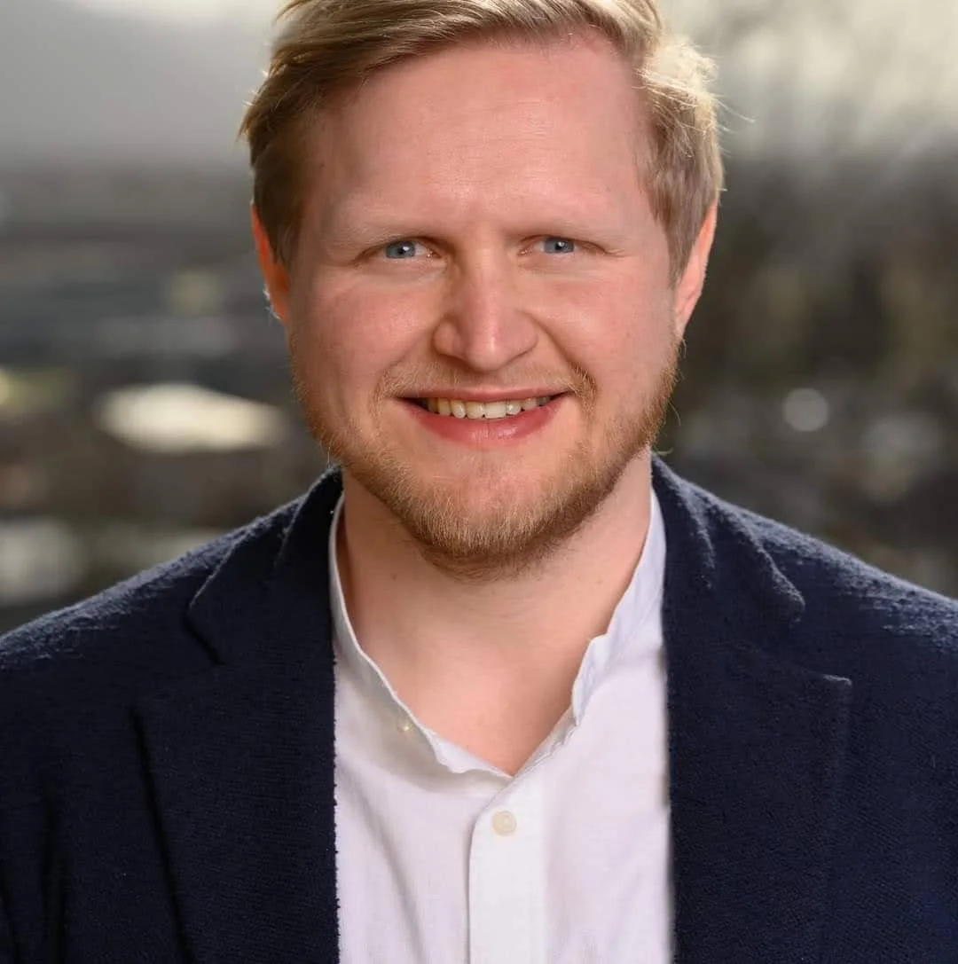 Ein Portrait eines lächelnden Mannes mit blonden Haaren und blauen Augen, trägt eine weiße Hemd und einen dunklen Blazer. Der Hintergrund ist unscharf mit natürlichen Lichtverhältnissen.