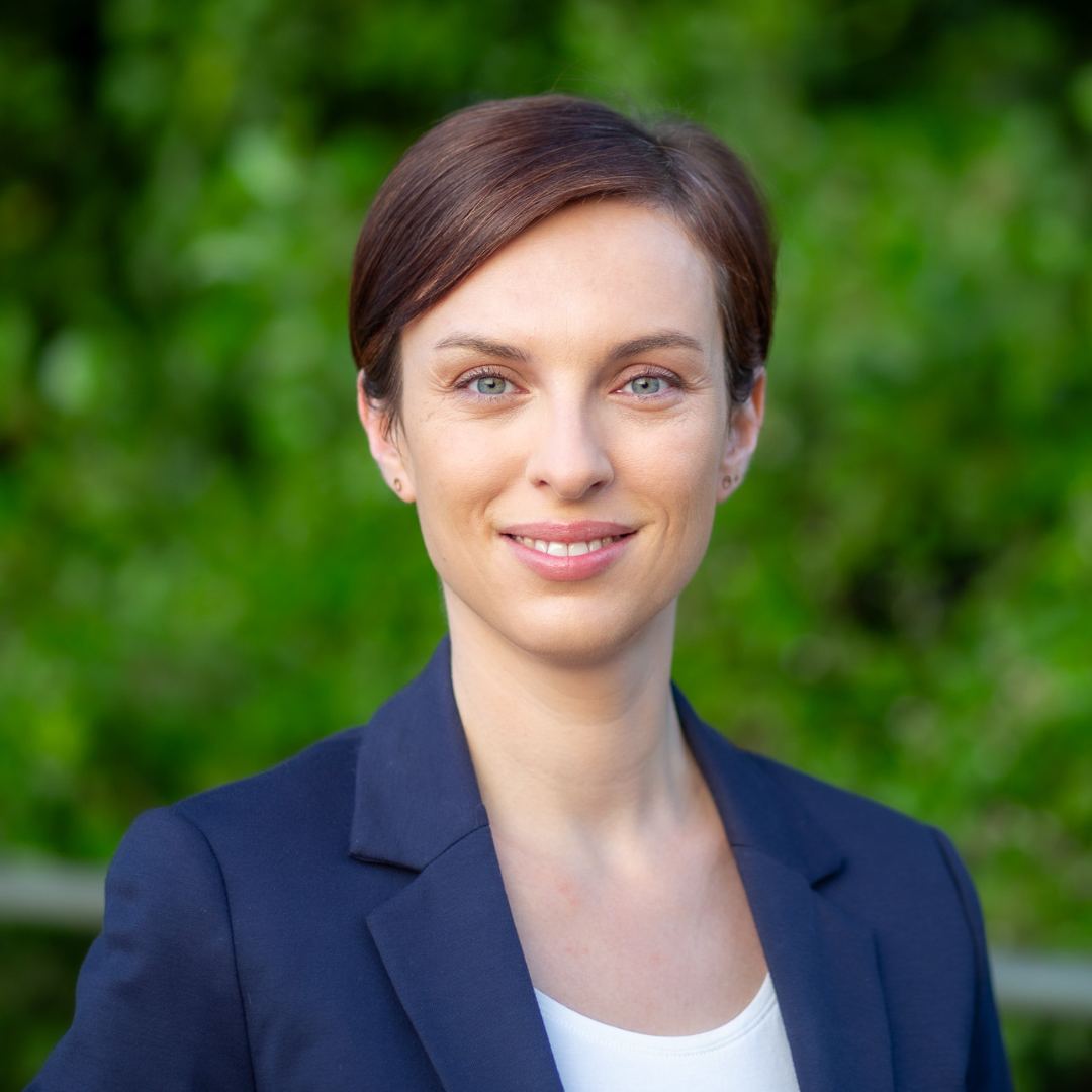 Porträt einer Frau mit braunen Haaren und blauen Augen, trägt einen dunkelblauen Blazer, vor grünem, naturbelassenem Hintergrund.