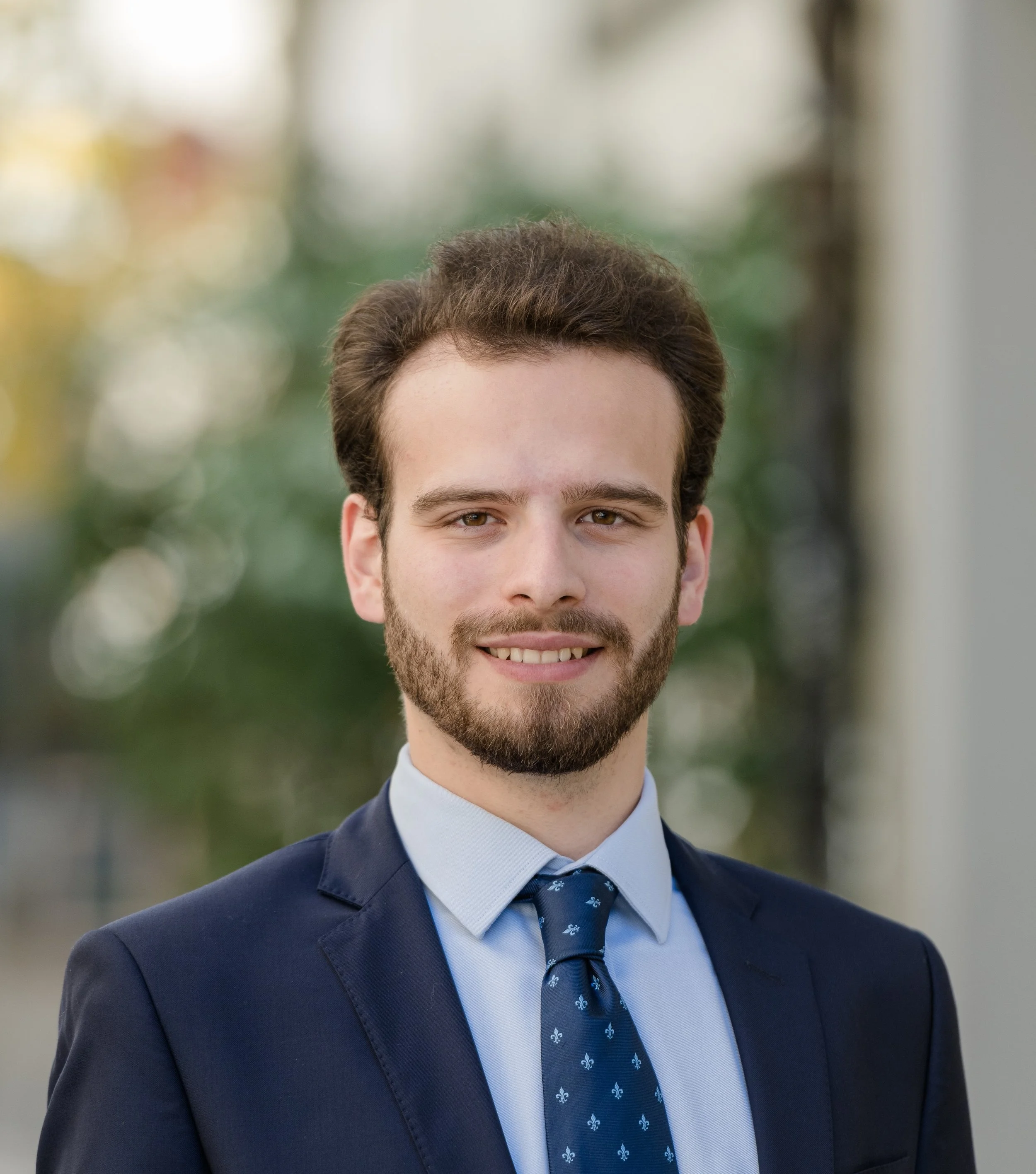 Der Mann trägt einen dunklen Anzug, weißes Hemd und eine blaue Krawatte mit kleinen weißen Mustern, steht draußen vor einem unscharfen Hintergrund mit Bäumen und Gebäuden.