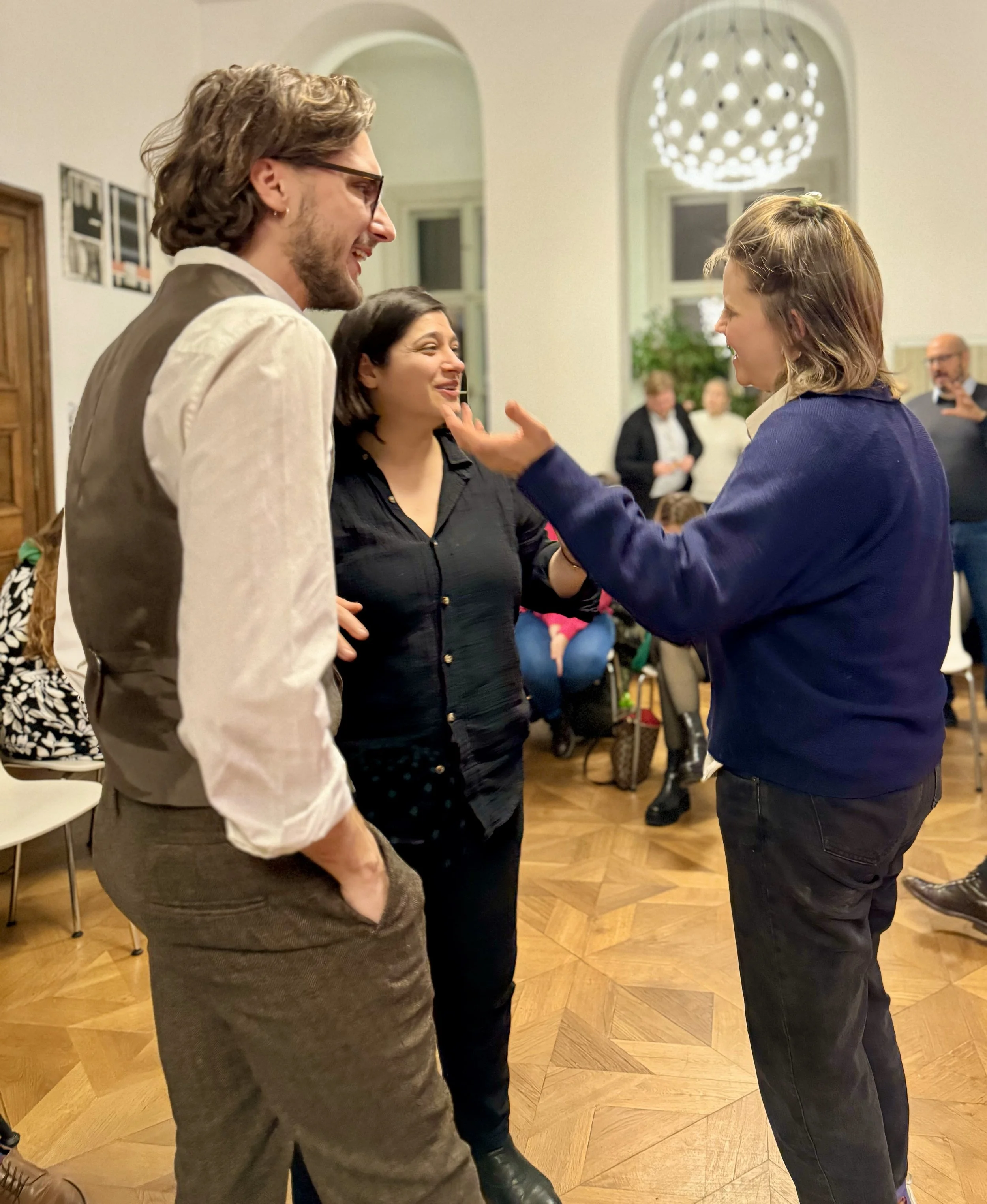 Drei Menschen unterhalten sich lachend in einem Wohnzimmer, weitere Personen im Hintergrund.