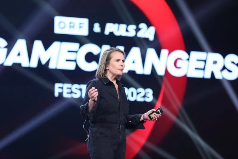 Eine Frau steht auf einer Bühne während eines Events namens GAMECHANGERS FESTIVAL 2023, im Hintergrund ein großes Display mit dem Text ORF & PULS 24 und weiteren Elementen.