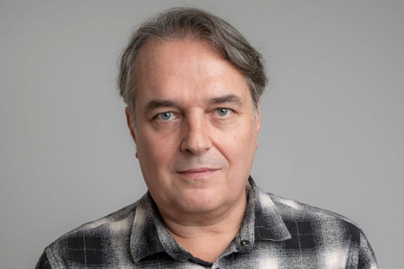 Porträt eines Mannes mit grauen Haaren, blauen Augen und einem schwarzen-weißen karierten Hemd vor grauem Hintergrund.