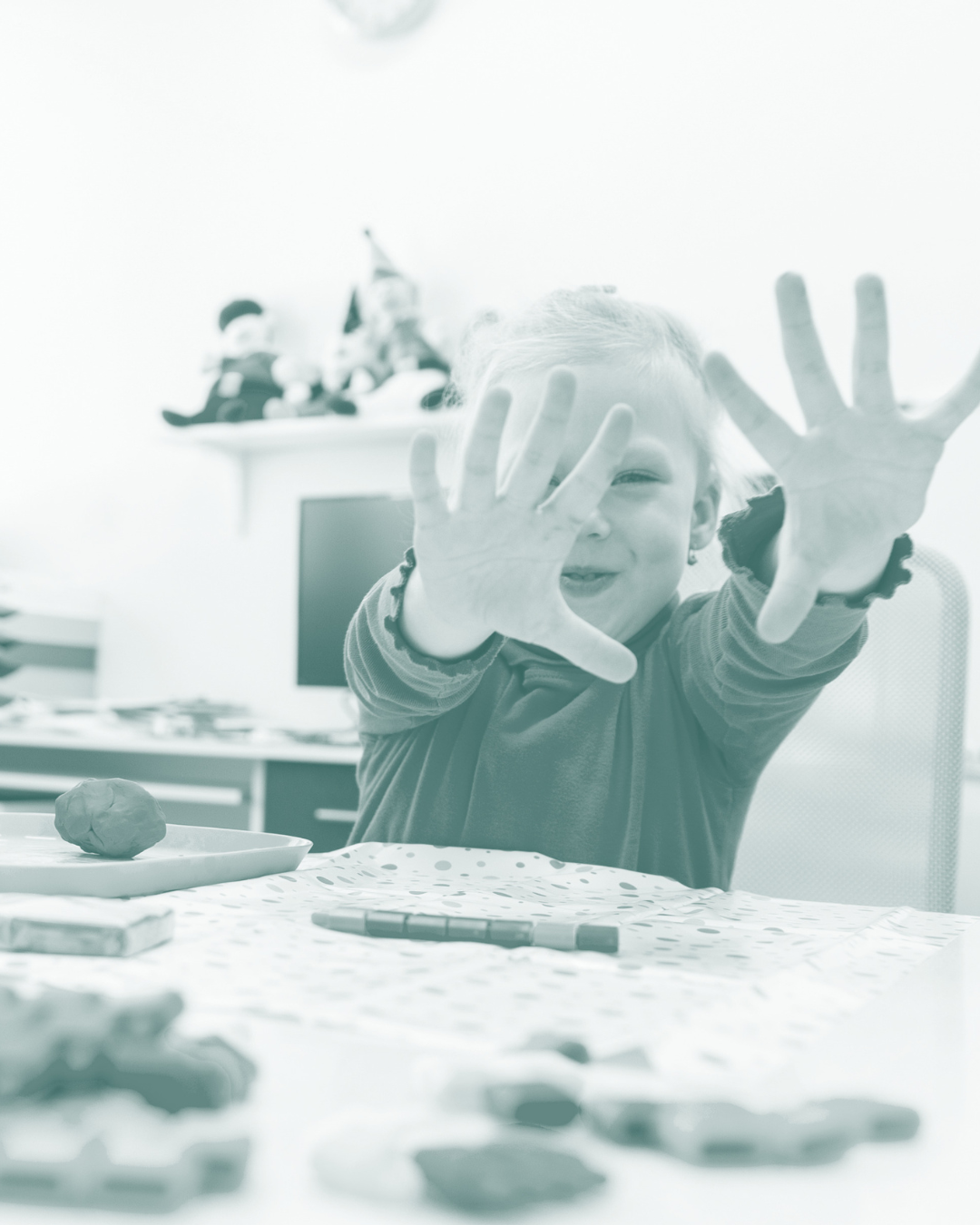A young child is playfully reaching out towards the camera with a big smile, sitting at a table covered with arts and crafts supplies and a piece of modeling clay in front of them. In the background, Christmas decorations are visible on a shelf.