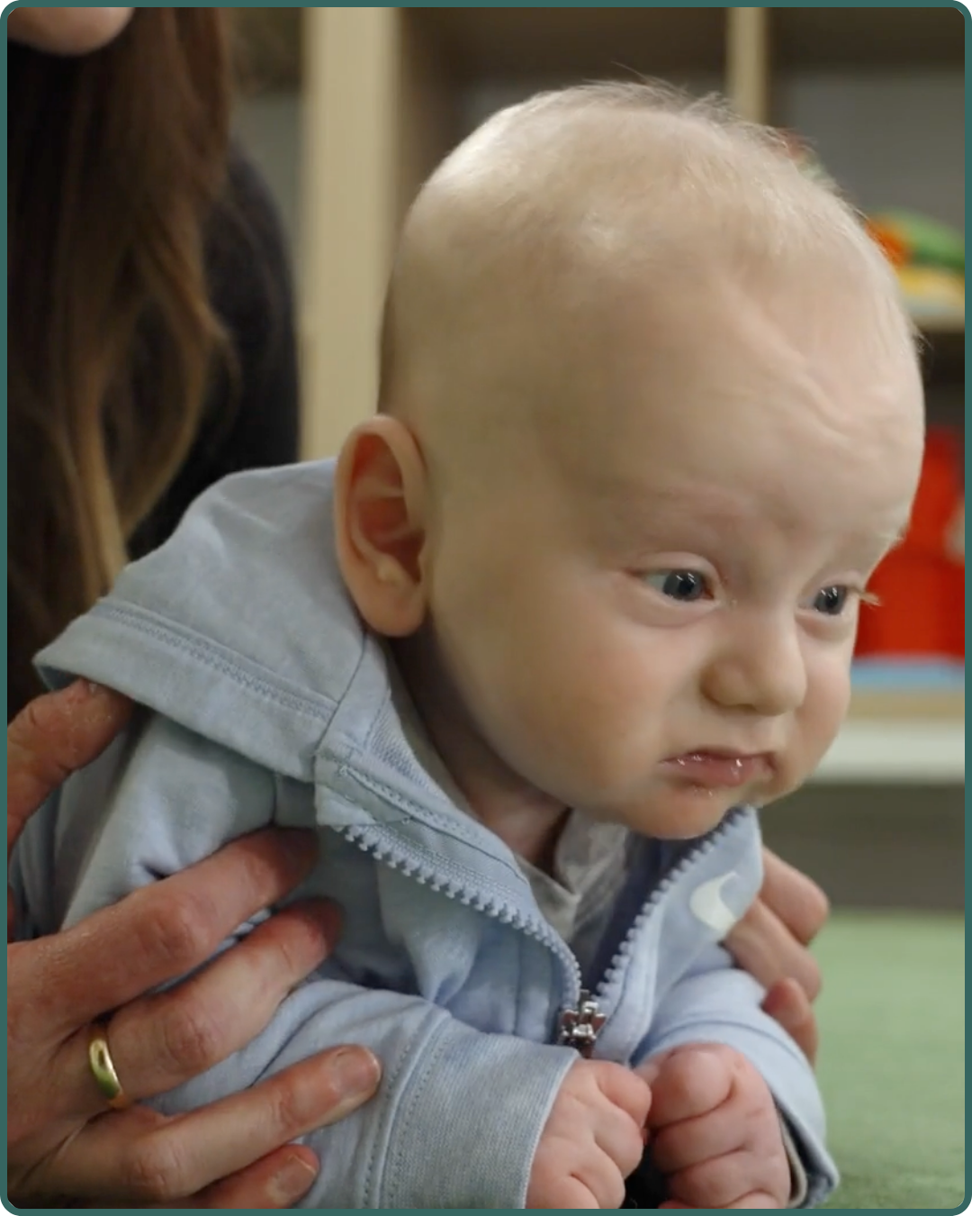 A baby with blonde hair and blue eyes in a gray hoodie, being supported by a person with a gold ring on their finger, in an indoor setting.