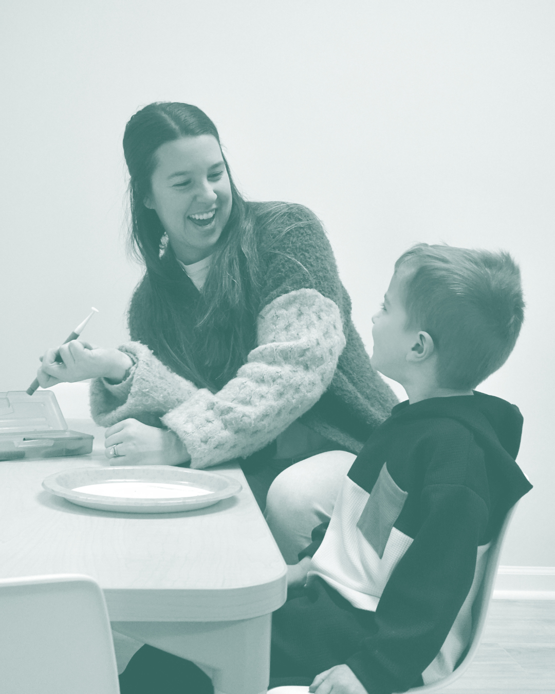 A woman smiling and talking to a young boy sitting at a table with a plate, holding a pen, in a simple room with plain white walls.