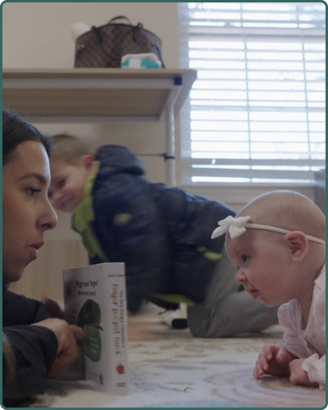 A woman and a baby girl lying on the floor facing each other, with a boy in the background, in a room with closed blinds and a table with a handbag on it.