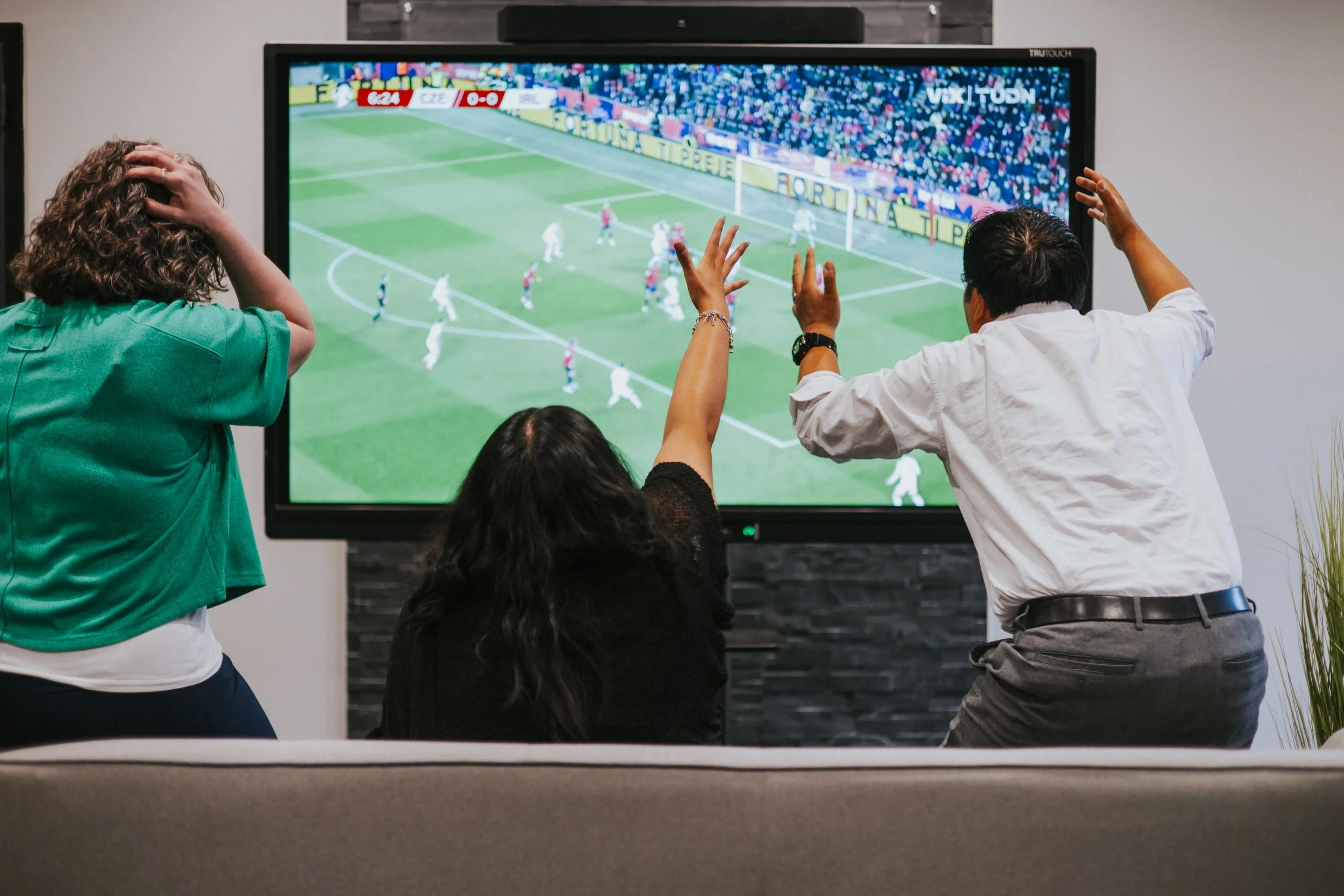 Three individuals watching a soccer match on a large television.