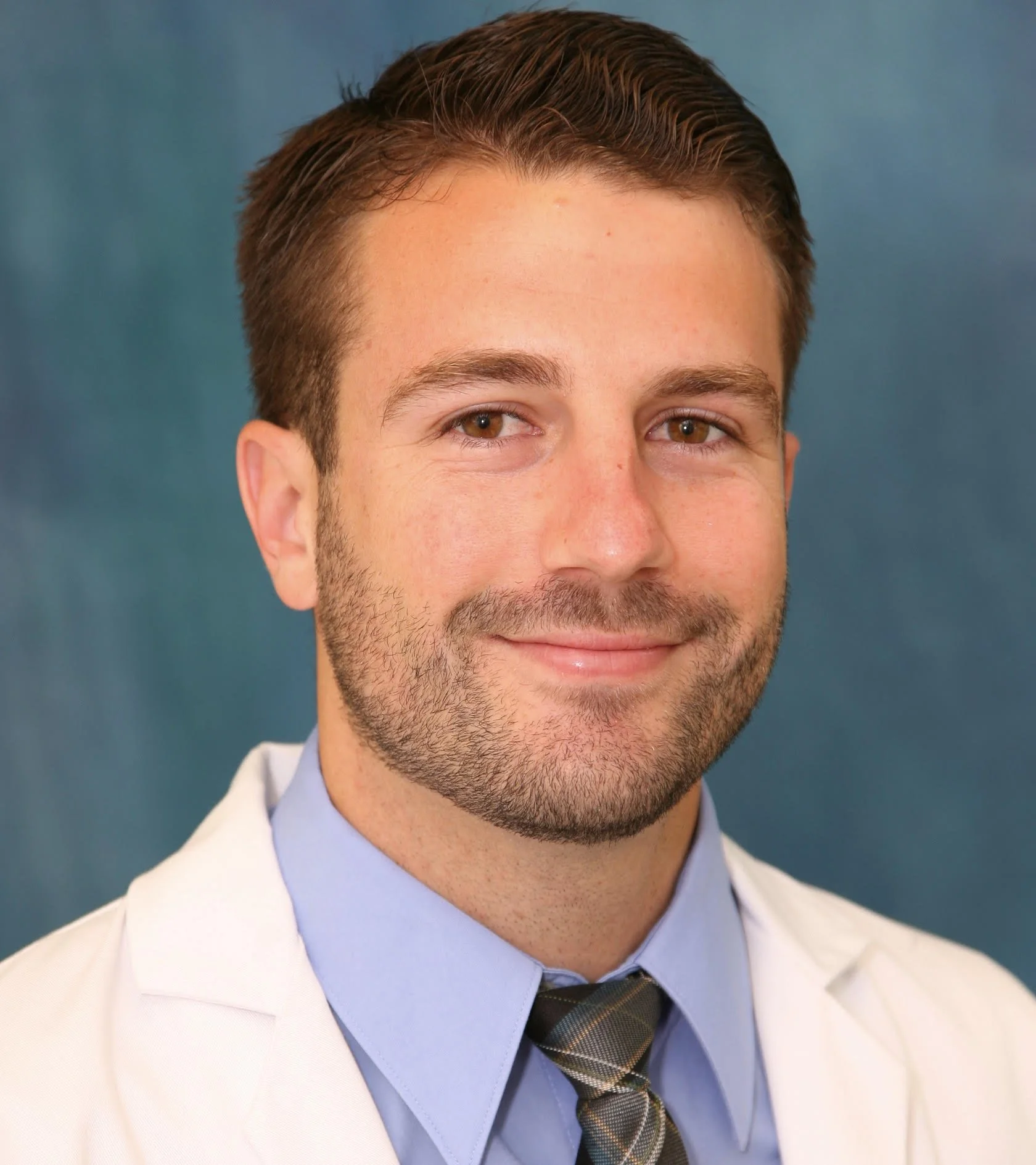 Close-up of a smiling man in a light-colored suit, blue shirt, and plaid tie, with short brown hair and a well-groomed beard, against a blue background.