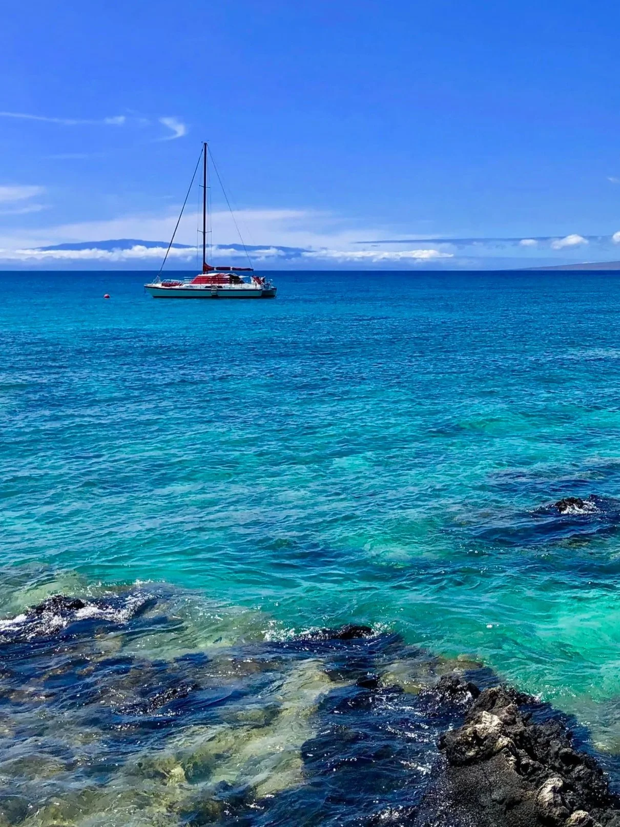A sailboat floating on turquoise ocean water near rocky shore under blue sky.