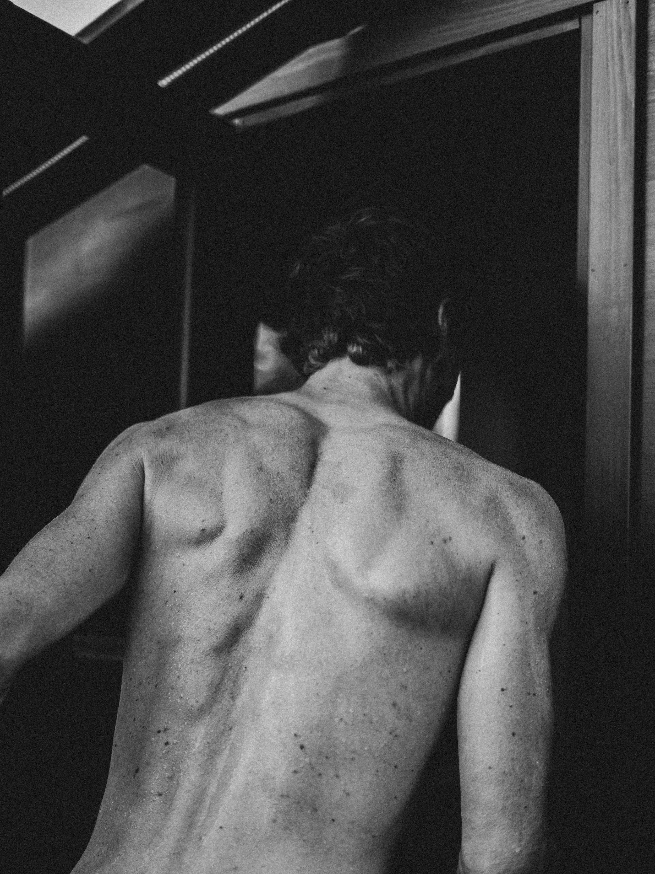 Vue en noir et blanc du dos d'une personne nue, le dos tourné, dans un environnement intérieur