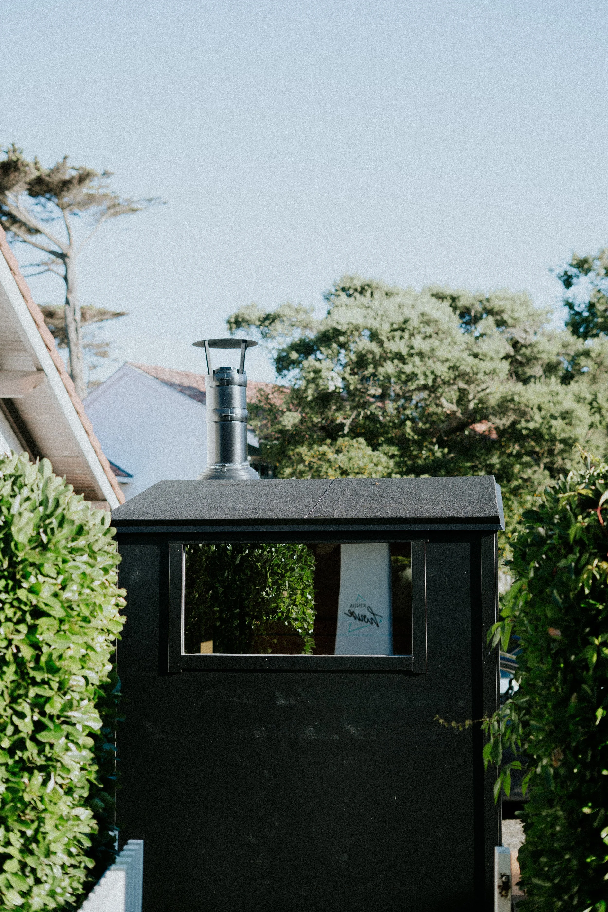 Un meuble noir avec une fenêtre et un panneau à l'intérieur, entouré de haies vertes, avec un ciel clair et des arbres en fond.