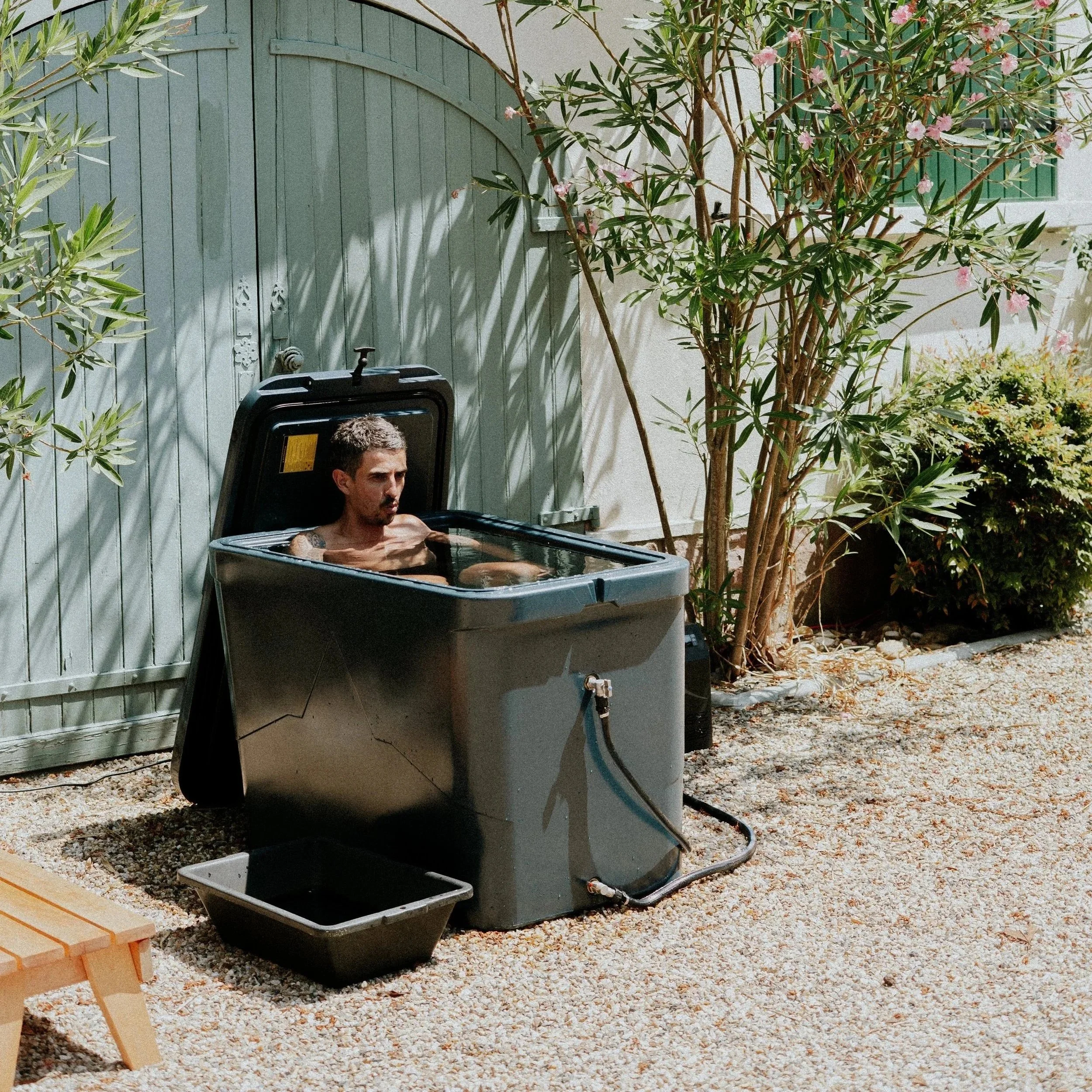 Un homme bronze dans une baignoire à bateau transformée en jacuzzi, située dans un jardin avec des plantes et un mur en bois vert.