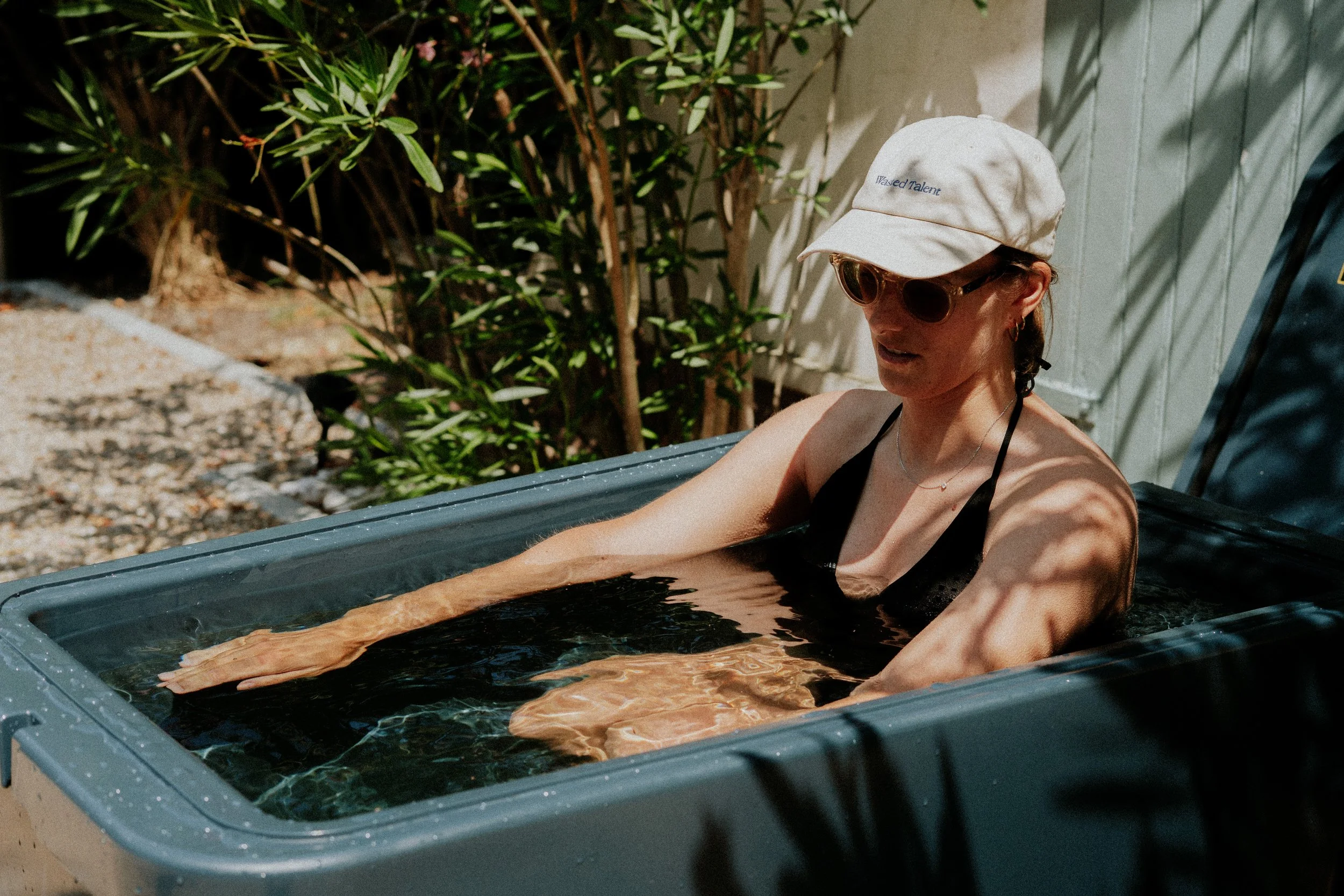 Une femme portant un chapeau blanc, des lunettes de soleil et un maillot de bain noir, se détend dans une baignoire d'eau noire en extérieur, avec des plantes en arrière-plan.
