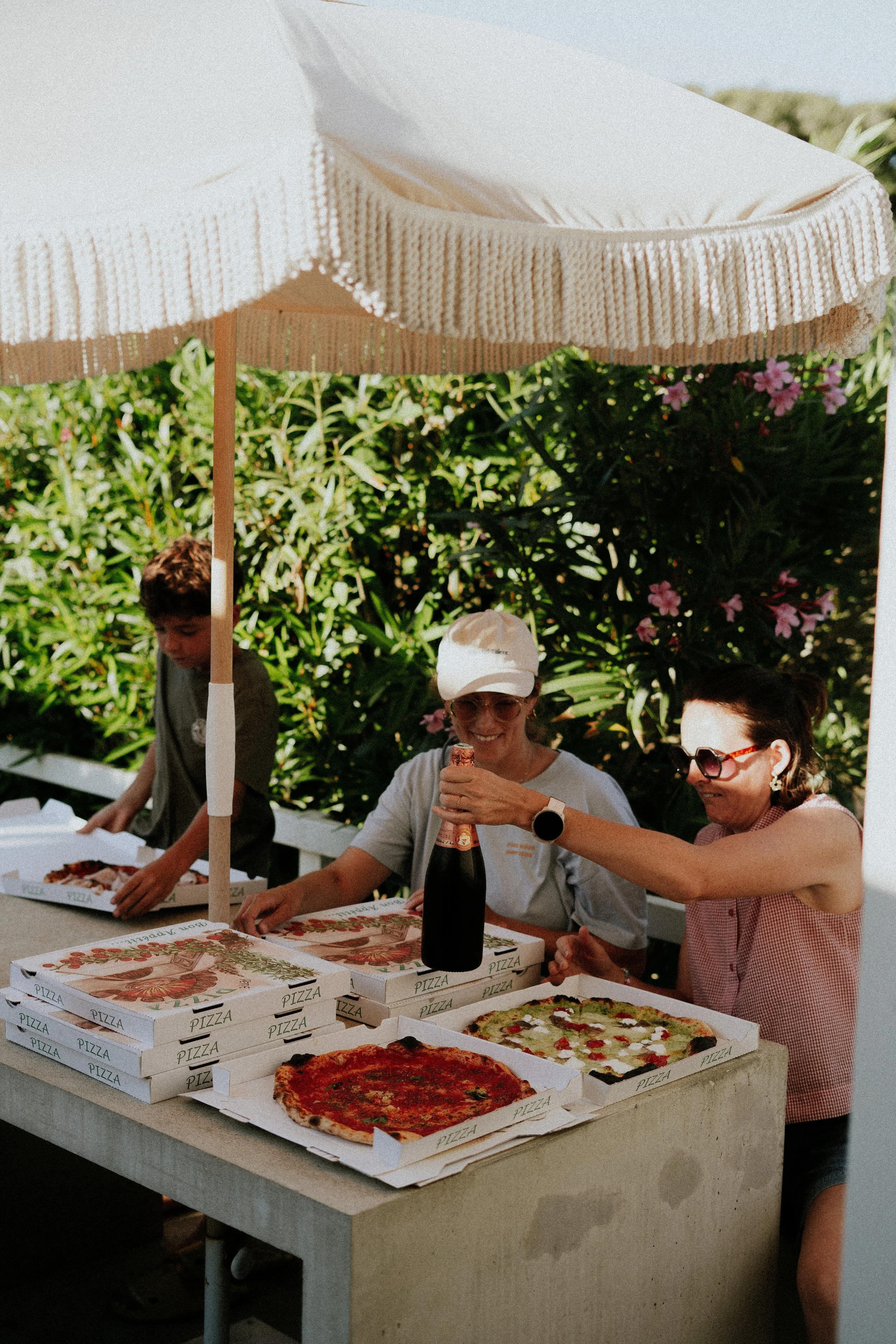 Des personnes assises autour d'une table avec des boîtes à pizza et une pizza en premier plan, sous un parasol blanc dans un jardin. Deux femmes portent des lunettes de soleil, une d'elles ouvre une bouteille.