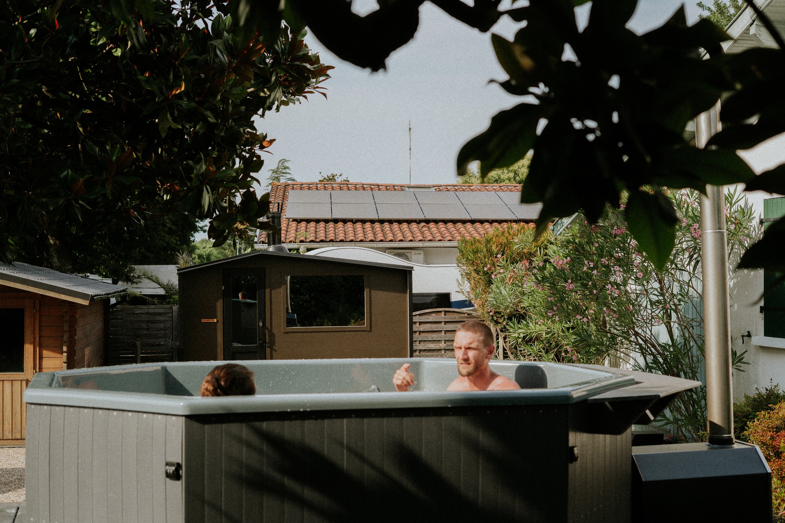 Deux personnes se détendent dans un jacuzzi dans un jardin entouré de végétation, avec une maison en arrière-plan ; le toit a des panneaux solaires.