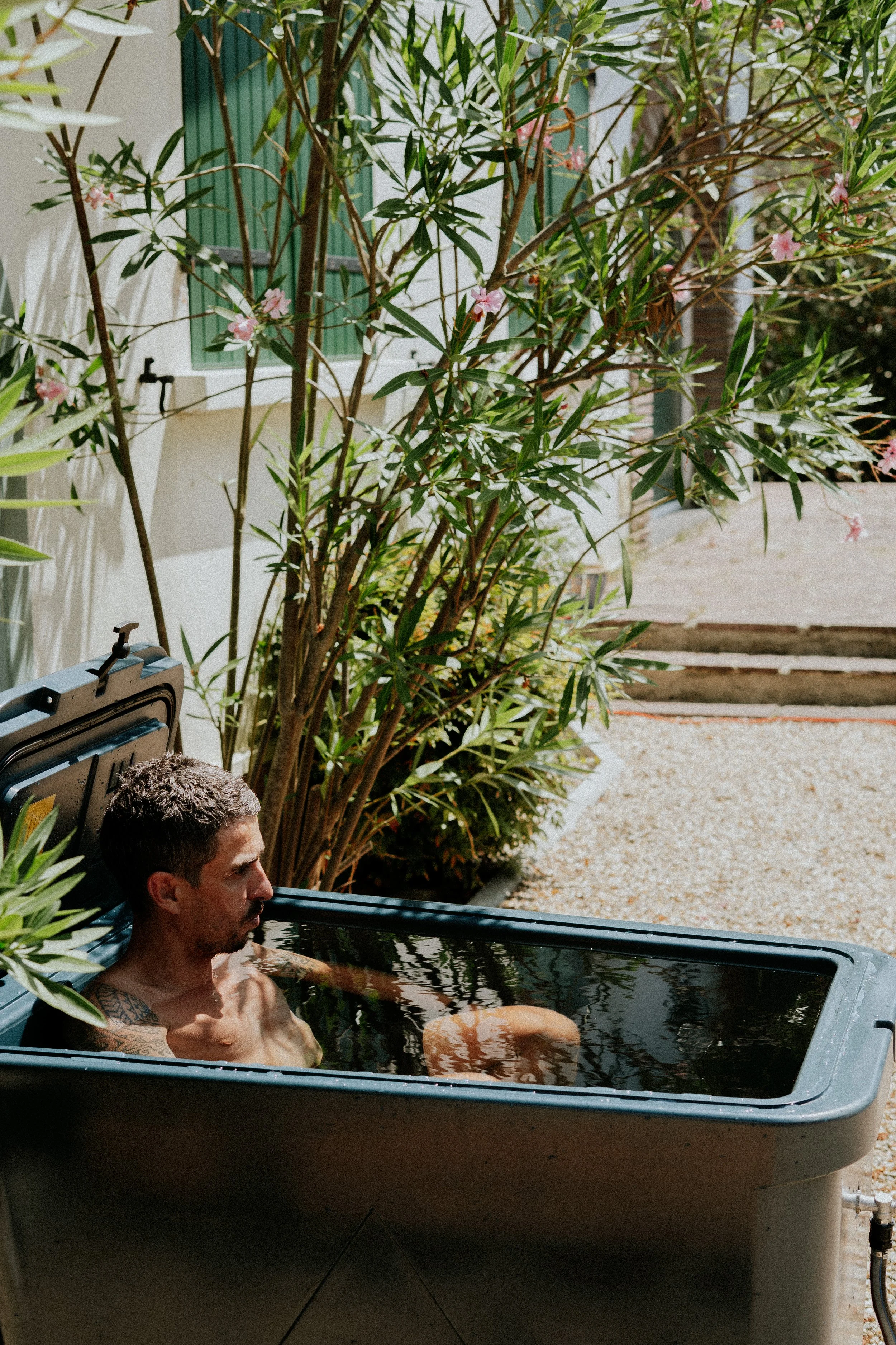 Un homme de tatouages se détend dans une baignoire extérieure remplie d'eau, entourée de plantes vertes, dans un espace en plein air ensoleillé.