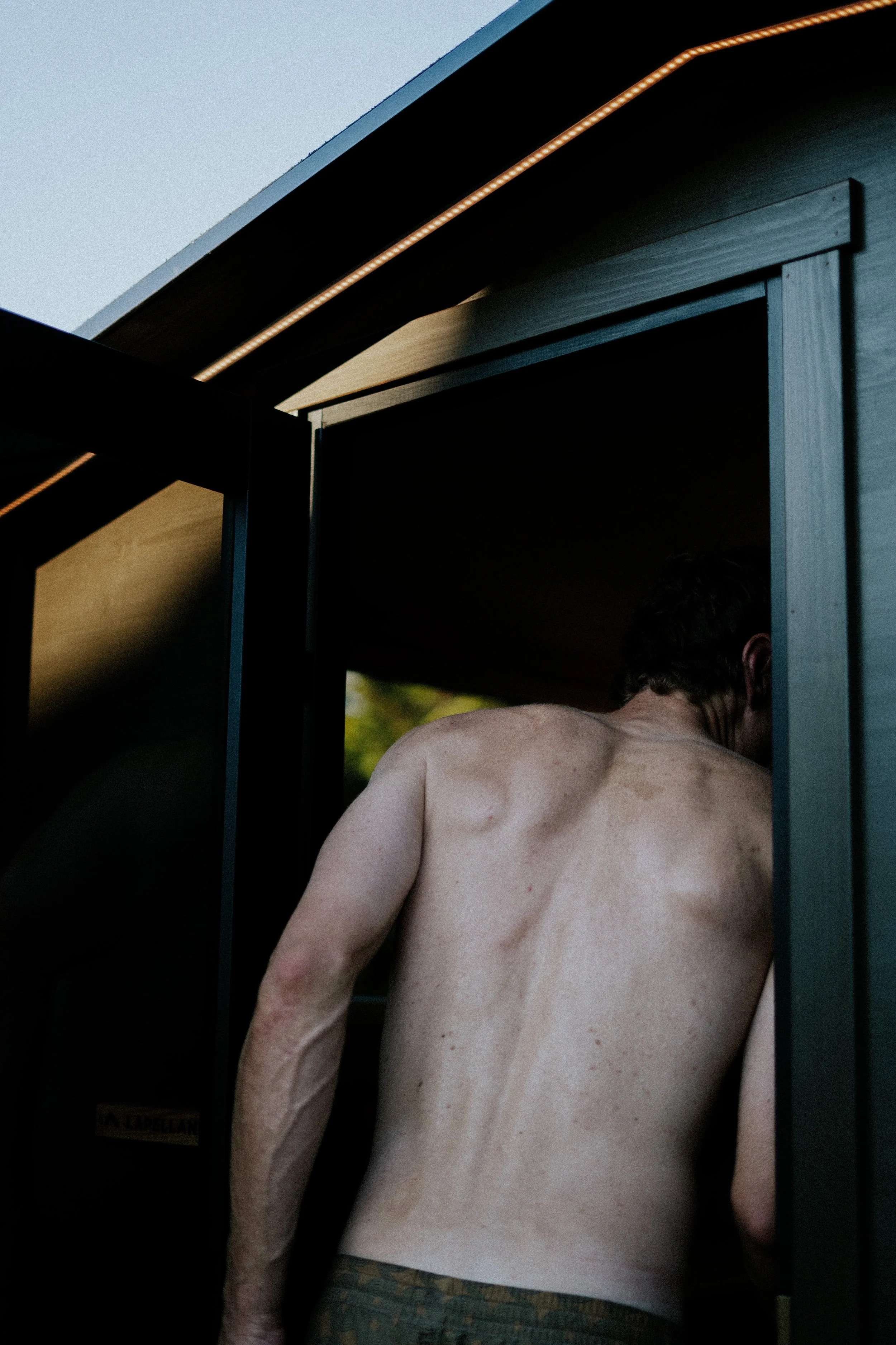 Un homme nu vu de derrière, avec peu de cheveux, en train de sortir d'une cabine ou d'un vestiaire.