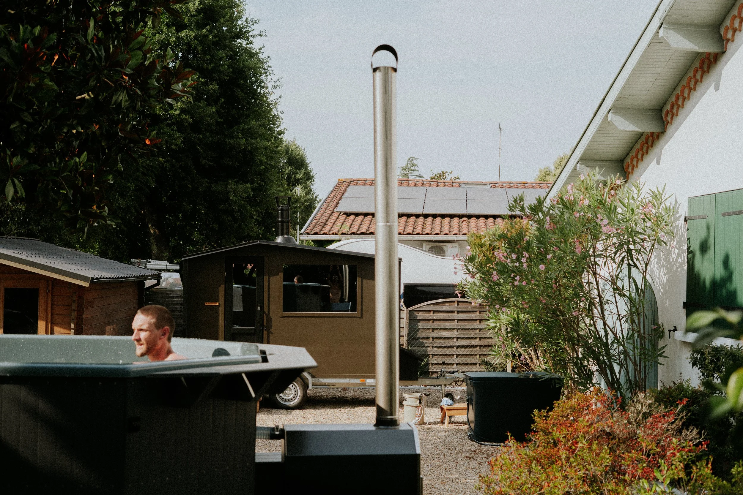 Une scène extérieure d'une cour avec un homme dans un jacuzzi, plusieurs plantes en pot, une maison avec un toit en tuiles, et un mur blanc avec une fenêtre, sous un ciel clair.