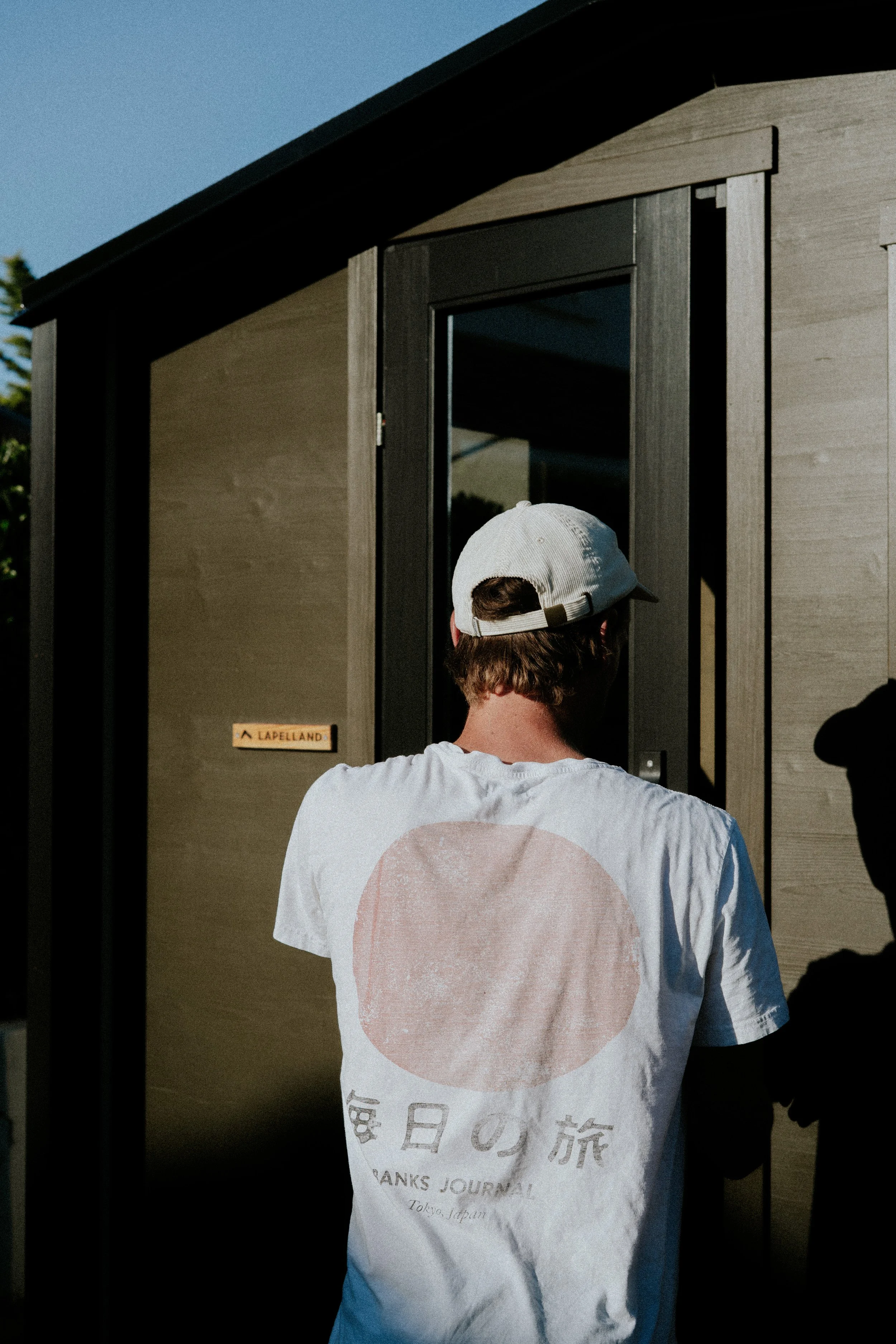 Un jeune homme portant un t-shirt blanc avec un design de cercle rose et des éléments de texte en japonais et anglais, une casquette beige, vu de dos, qui se tient devant une porte en verre d'une structure en bois beige, sous un ciel clair.