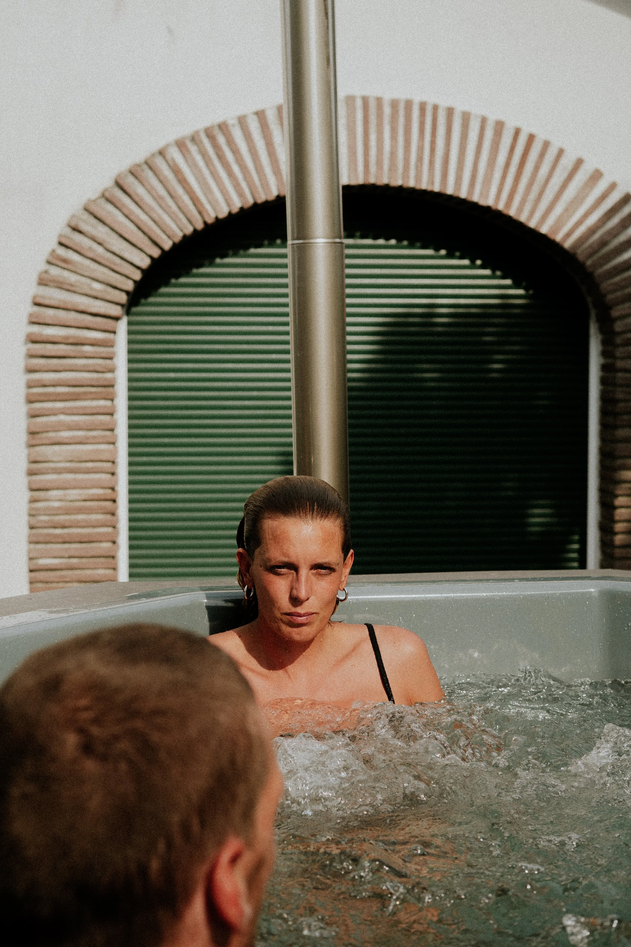Une femme d'apparence déterminée, avec des cheveux sombres attachés en arrière, est dans une piscine ou un jacuzzi, vue partielle en gros plan, avec une paroi verte en arrière-plan et une arche de briques à gauche.