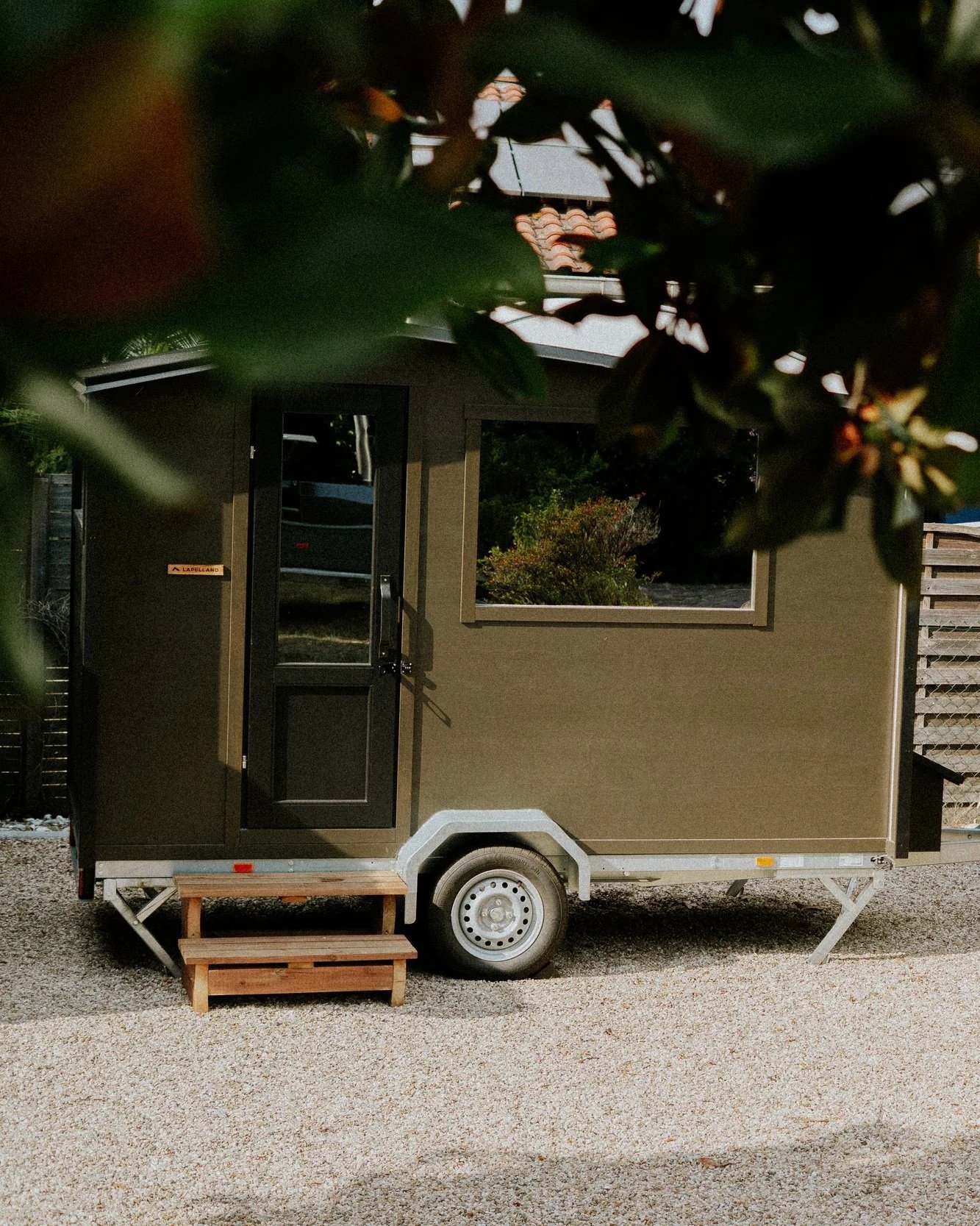 Une petite maison mobile noire avec une porte en verre et une fenêtre, placée sur un terrain en gravier, entourée d'arbres et de végétation, avec un petit escalier en bois devant la porte.