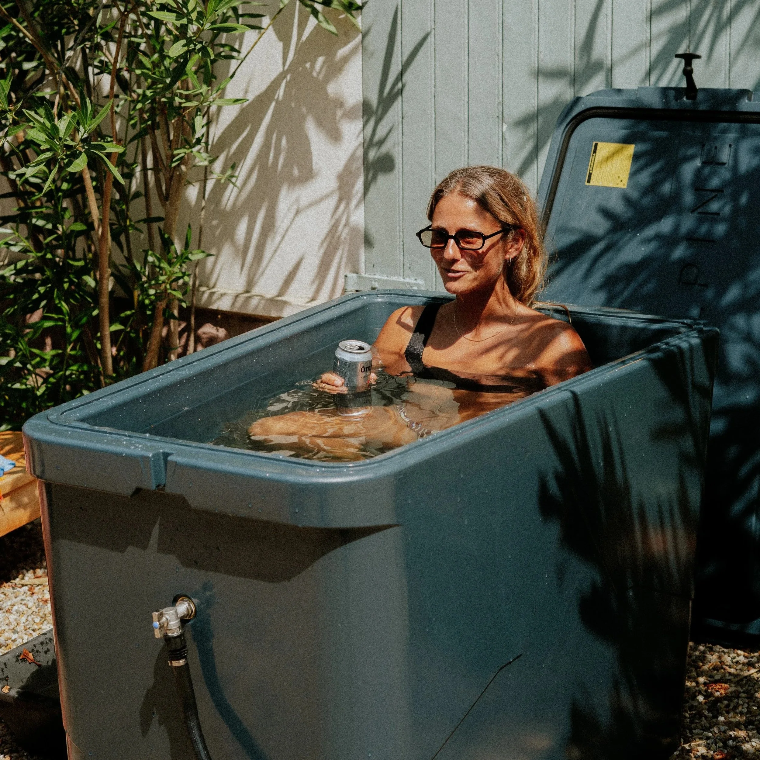 Une femme assise dans une poubelle bleue, tenant une canette de boisson. Elle porte des lunettes et un débardeur noir, avec des lunettes de soleil sur la tête. Il y a un mur en arrière-plan, une plante à gauche, et l'ombre de feuilles sur le mur.