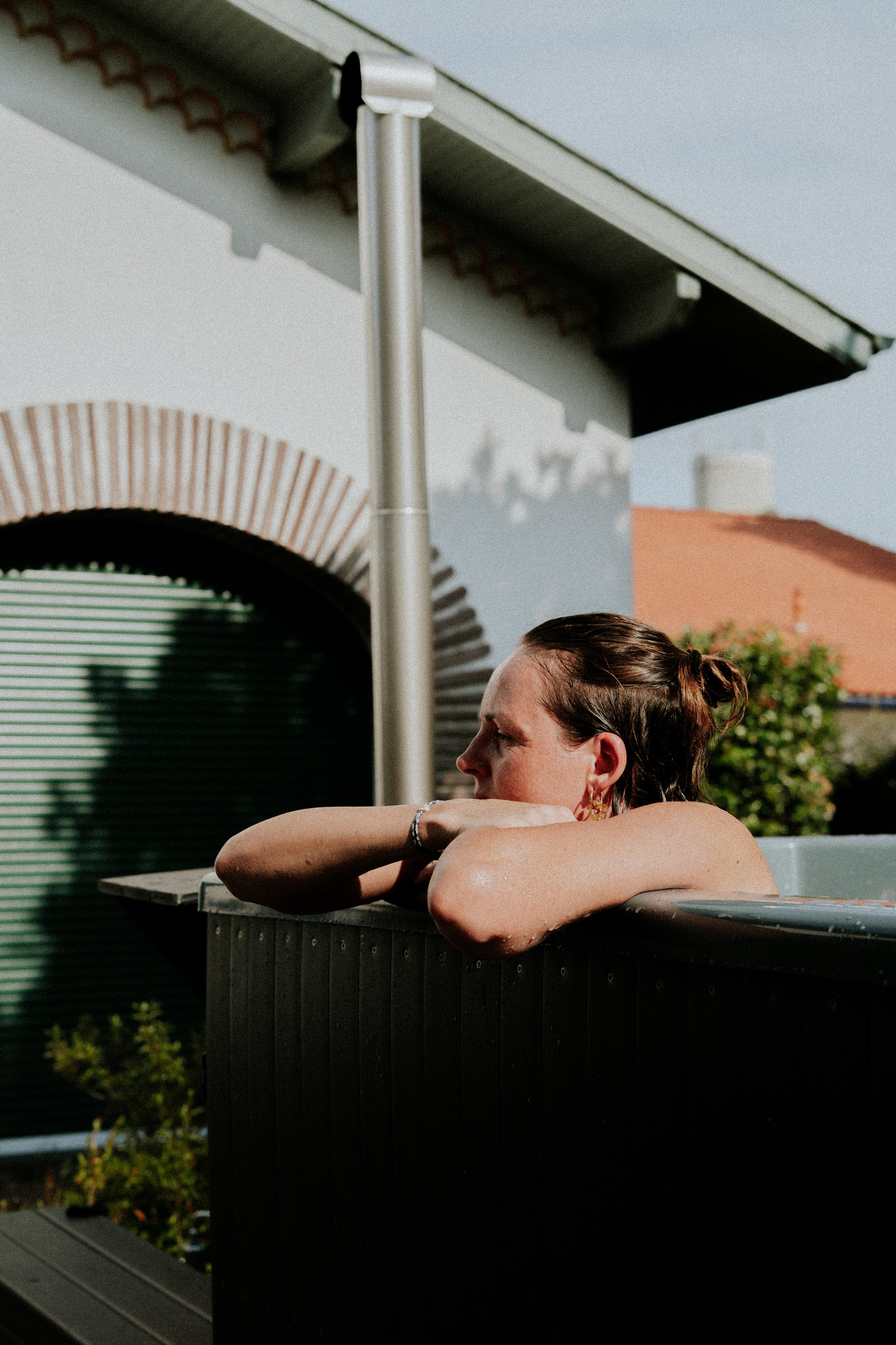 Une femme avec des cheveux bruns attachés regarde vers la droite, appuyée sur le rebord d'une piscine ou d'un jacuzzi en extérieur, avec un bâtiment coloré en arrière-plan, par une journée ensoleillée.