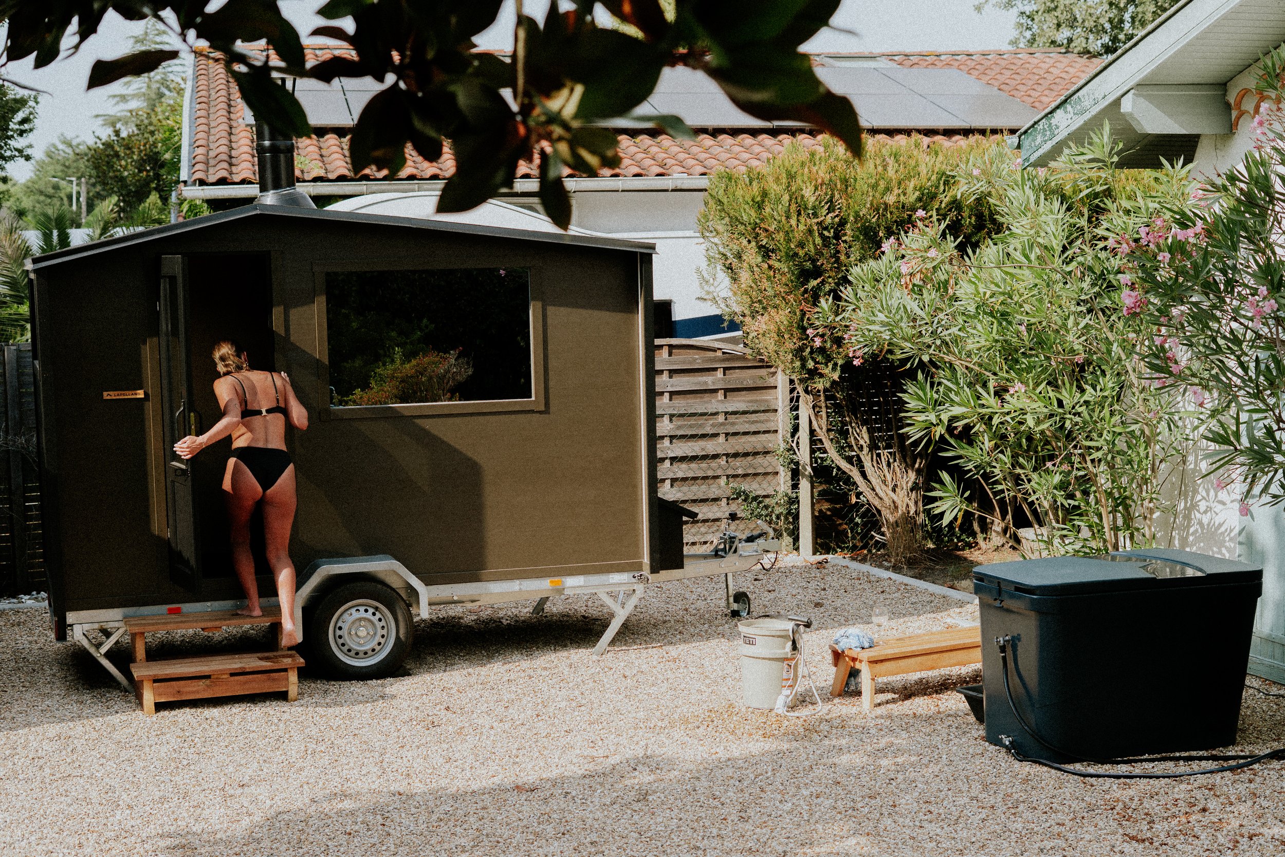 Une femme en maillot de bain noir quitte une tiny house sur roulettes dans une cour bordée d'arbres et de fleurs, avec une toiture en tuiles.