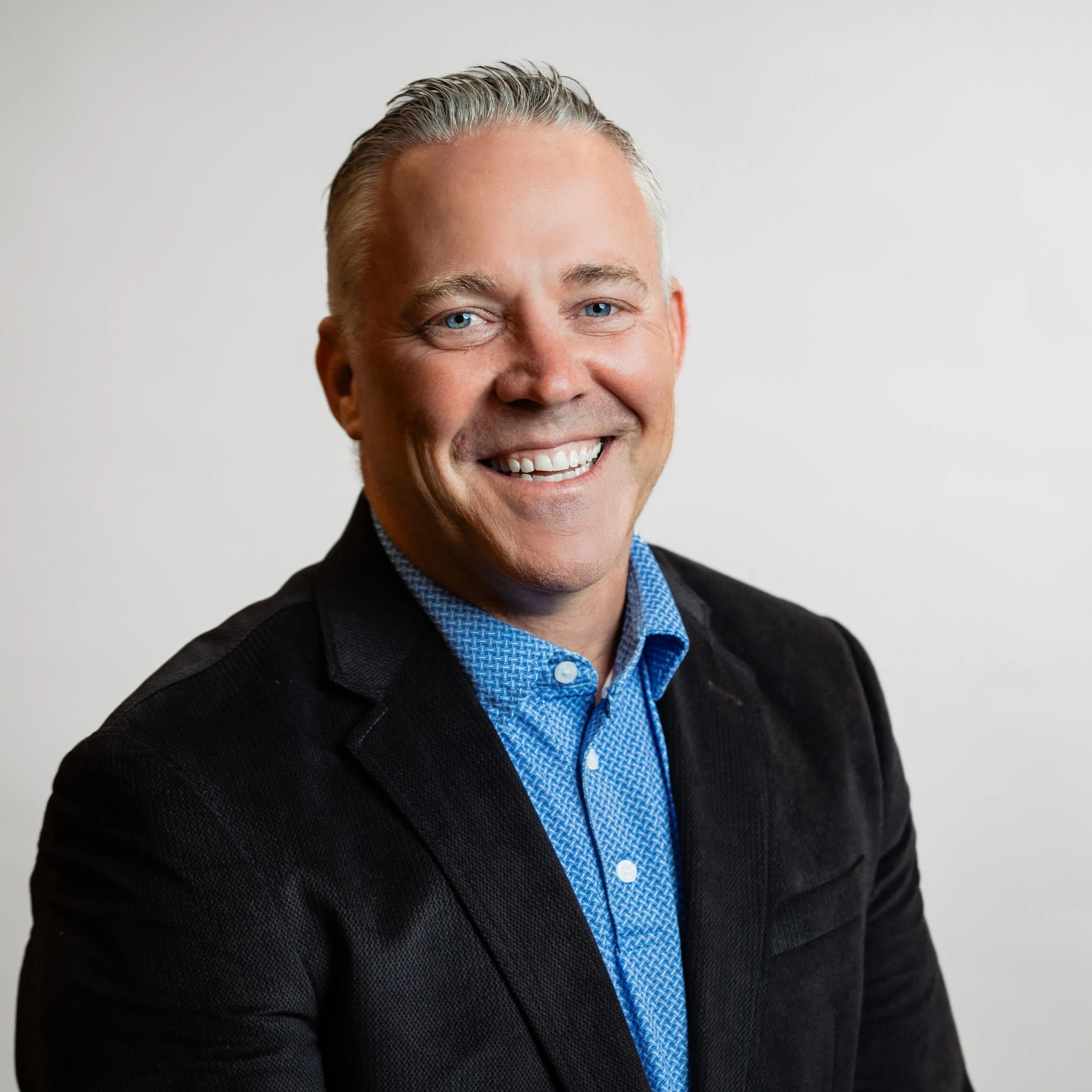 A smiling man with short, gray hair wearing a black blazer and a blue patterned button-up shirt against a plain white background.