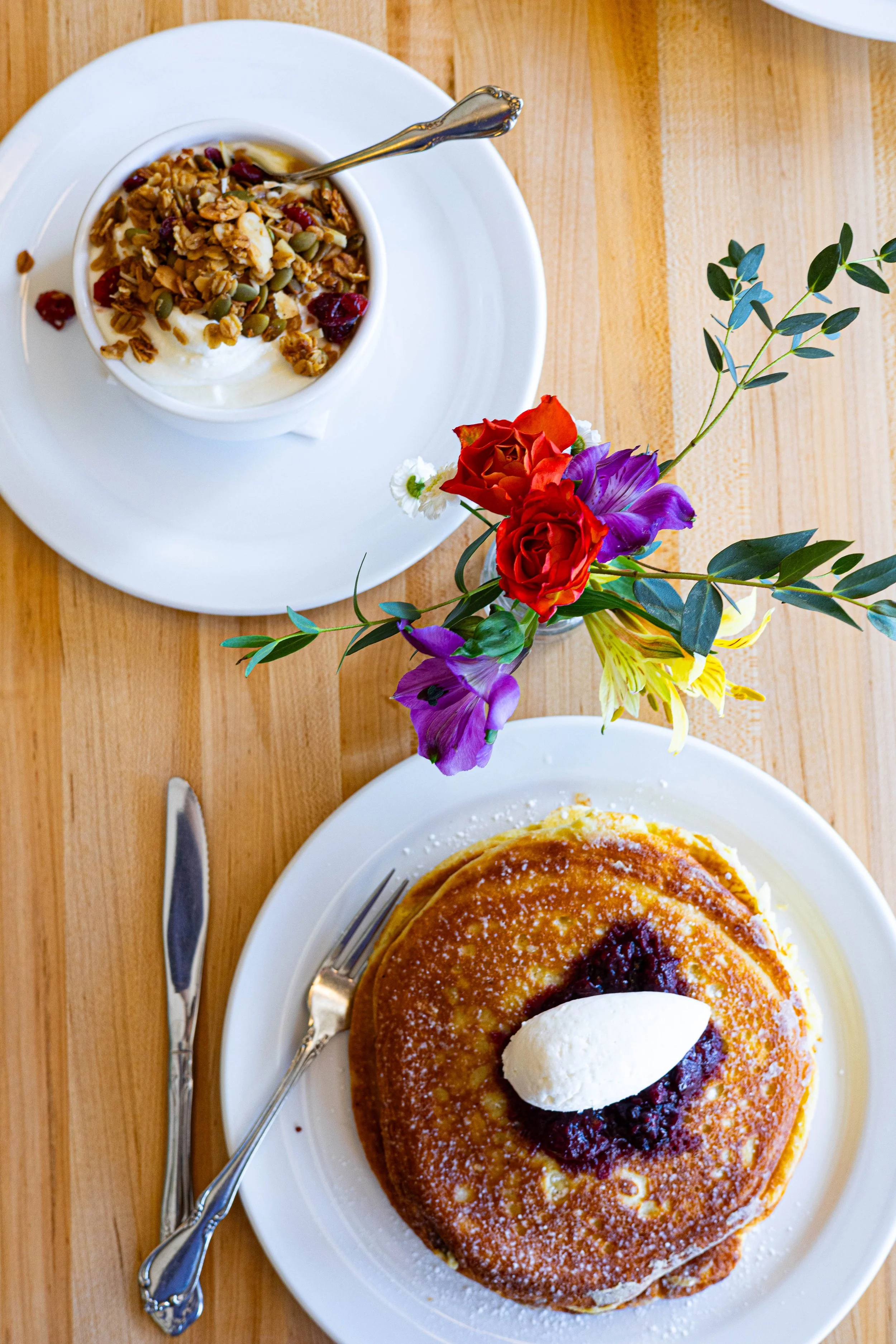 A breakfast setting with a plate of pancakes topped with a dollop of cream and fruit jam, a bowl of yogurt with granola and dried fruit, and a small bouquet of colorful flowers, all on a wooden table.