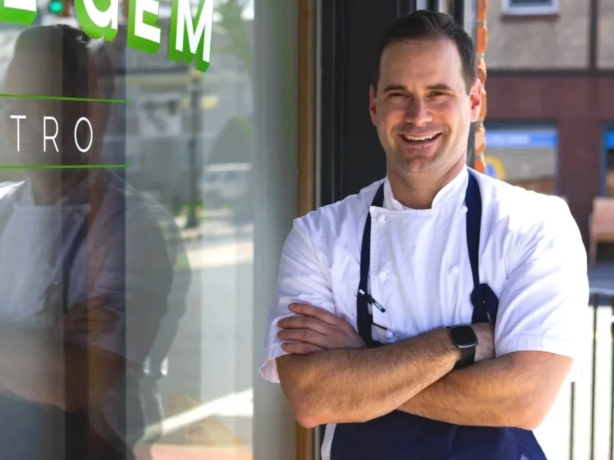 A smiling male chef in a white chef's coat and apron standing outside a building with a glass window, crossing his arms.