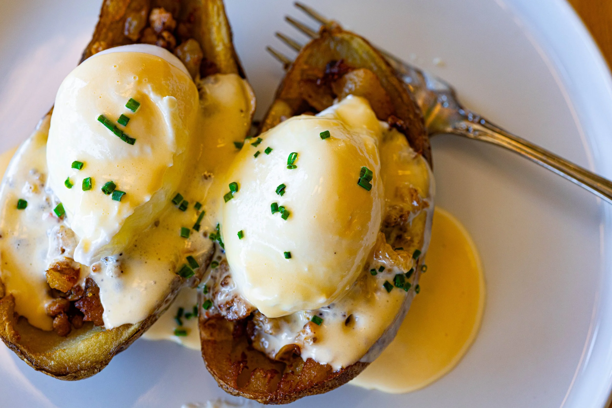 Baked potato halves topped with poached eggs, creamy sauce, and chopped chives on a white plate with a fork.