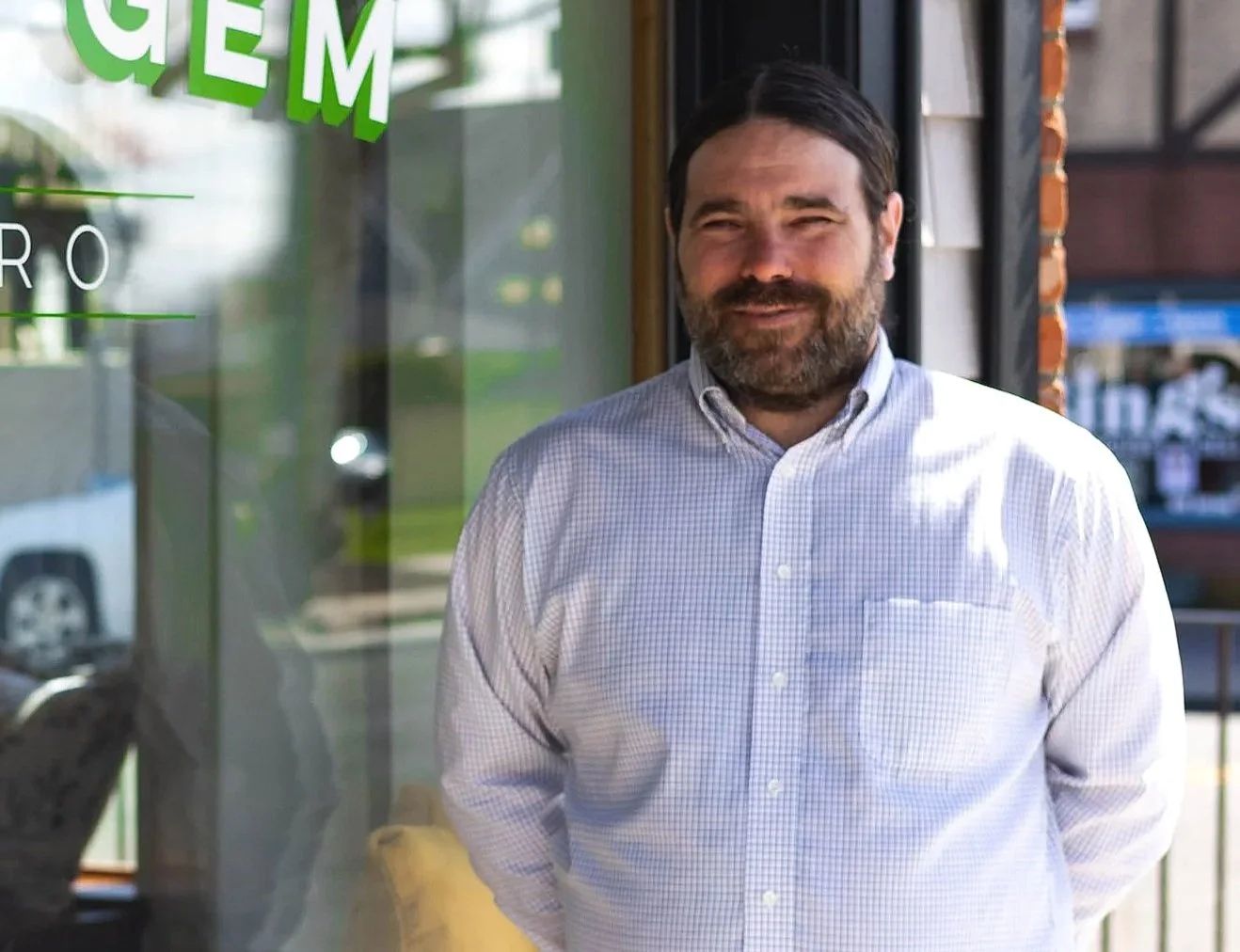 A man with dark hair and a beard wearing a light-colored, checkered button-up shirt standing outside a shop with a glass door window.