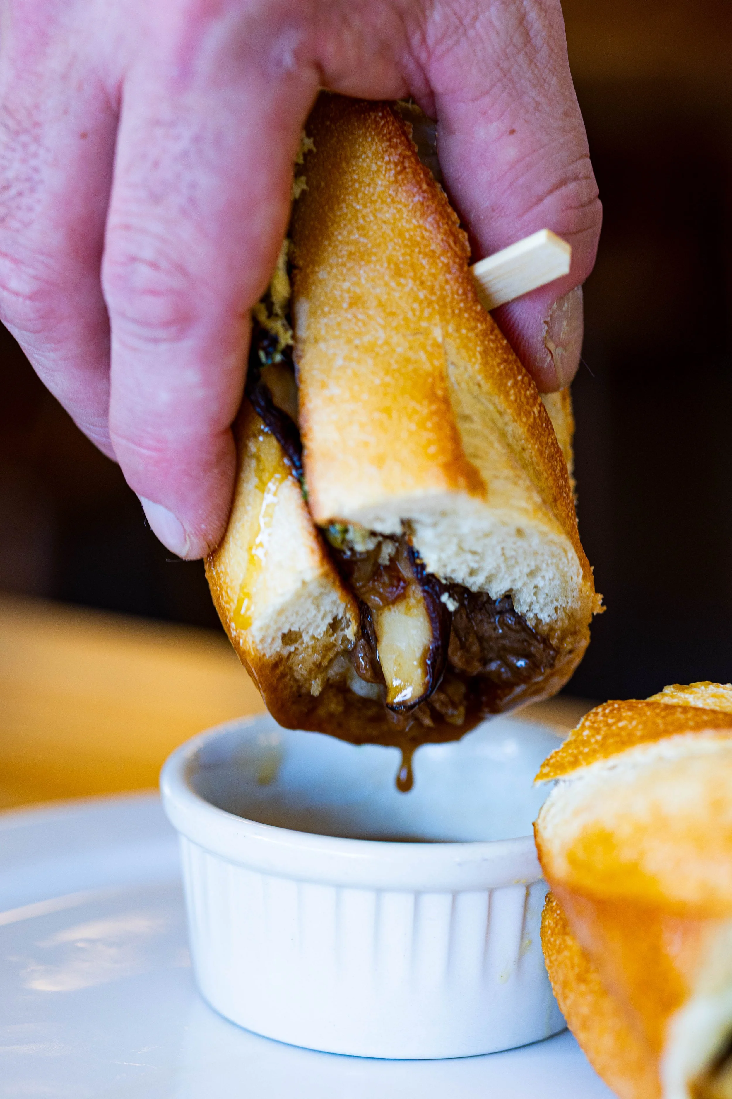 Close-up of a hand holding a toasted sandwich with melted chocolate inside over a small white bowl, with another toasted sandwich on a white plate in the foreground.