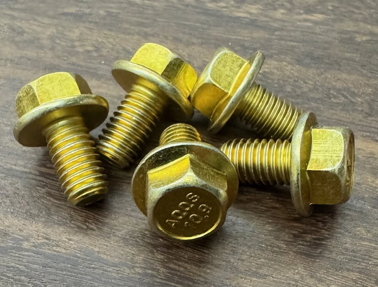 Six gold-colored metal bolts with hexagonal heads and threaded shafts are scattered on a wooden surface.