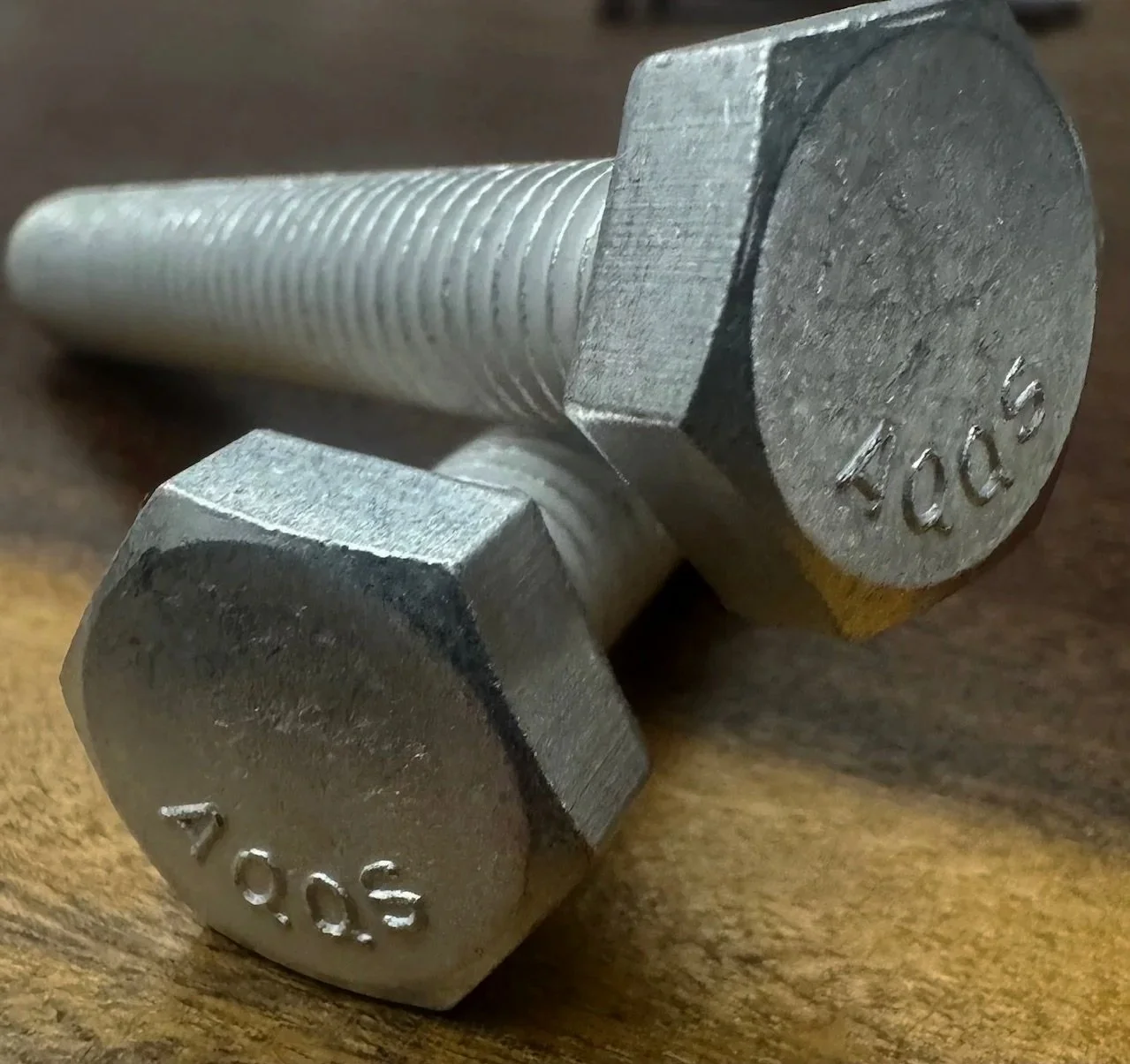 A close-up photo of two metallic hexagonal bolts on a wooden surface. The bolts have threaded shafts, and the heads are engraved with the markings "10.05" and "4.00".