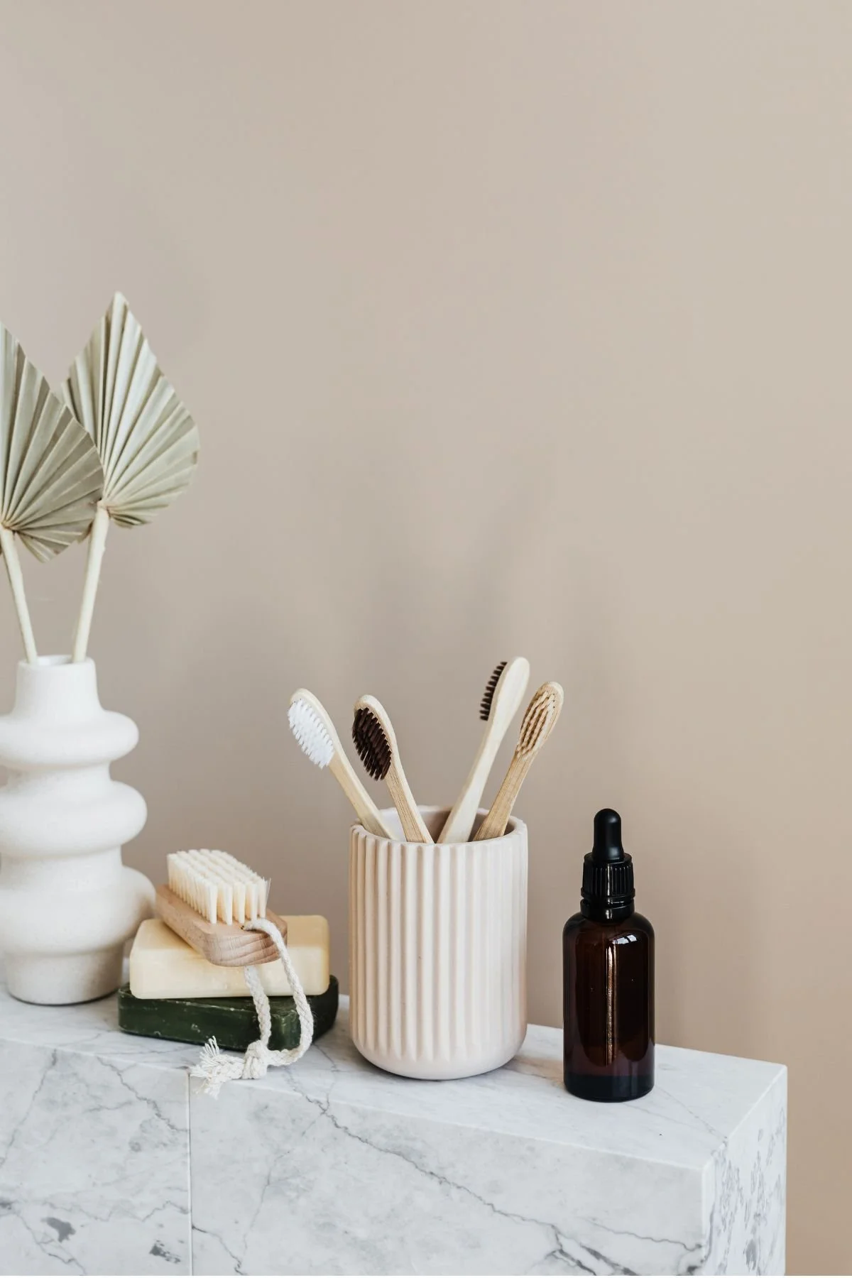 A marble countertop with candles, a soap bar, bamboo toothbrushes in a beige ribbed ceramic holder, a black dropper bottle, and dried palm leaves in a white vase.