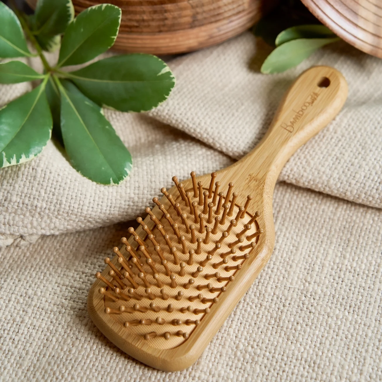 Wooden hairbrush with bristles on a beige fabric surface, with green leaves and woven baskets in the background.