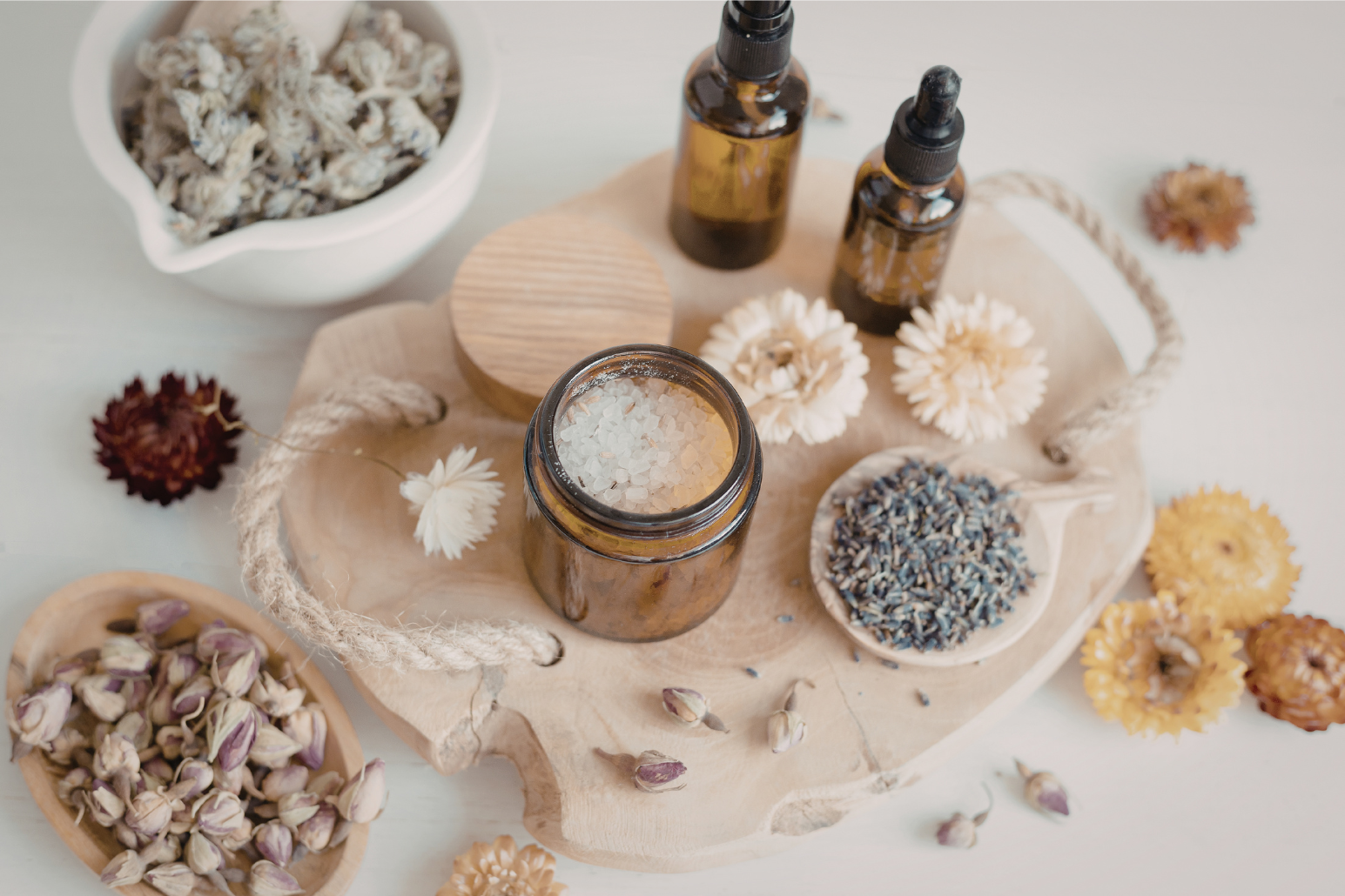 A wooden tray holds a jar of bath salts, two amber glass bottles with droppers, a bowl of lavender buds, and dried flowers. Additional dried flowers are scattered around.